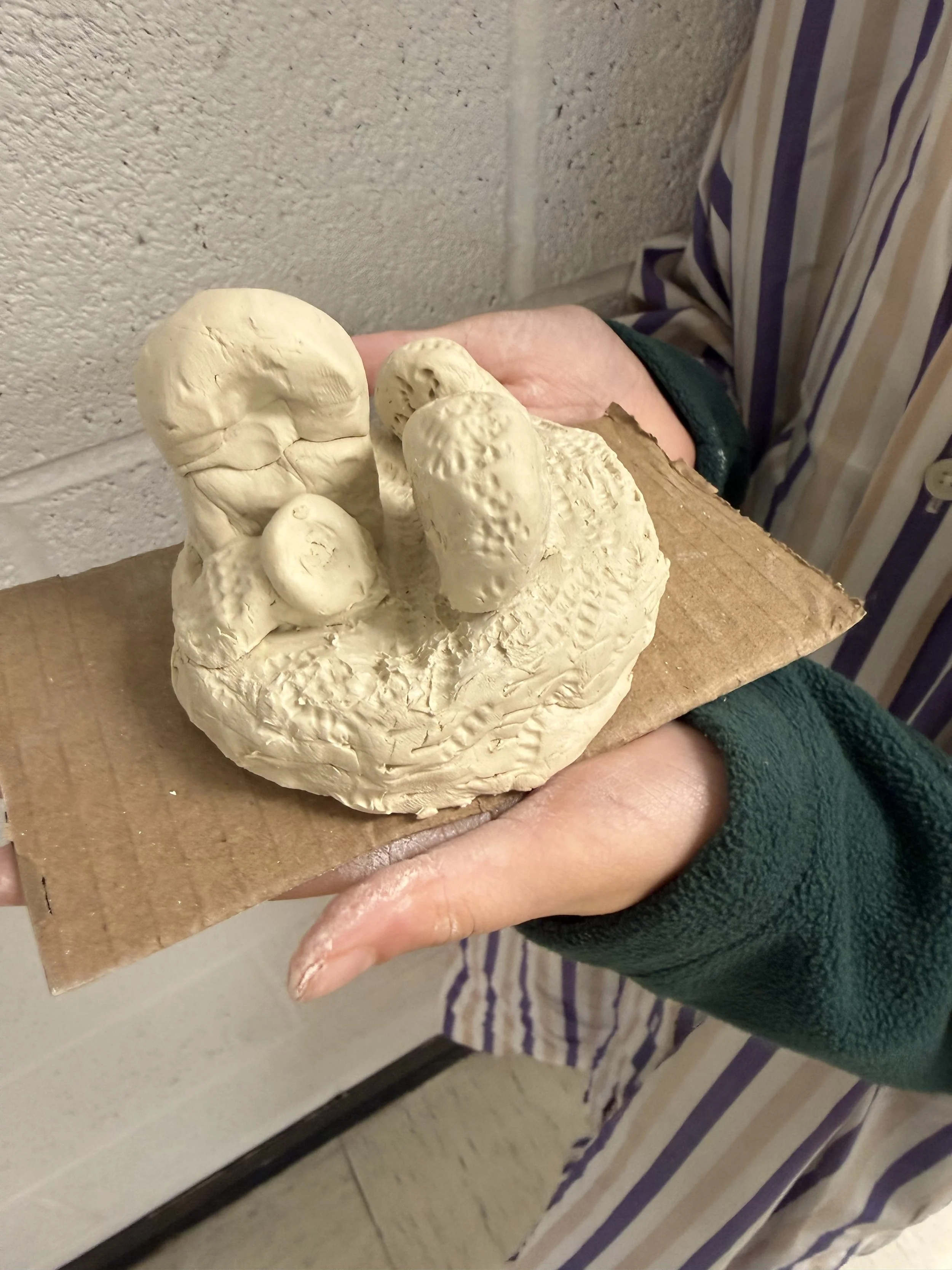 Hand holding a small clay sculpture on a piece of cardboard, depicting abstract shapes resembling animals or figures.