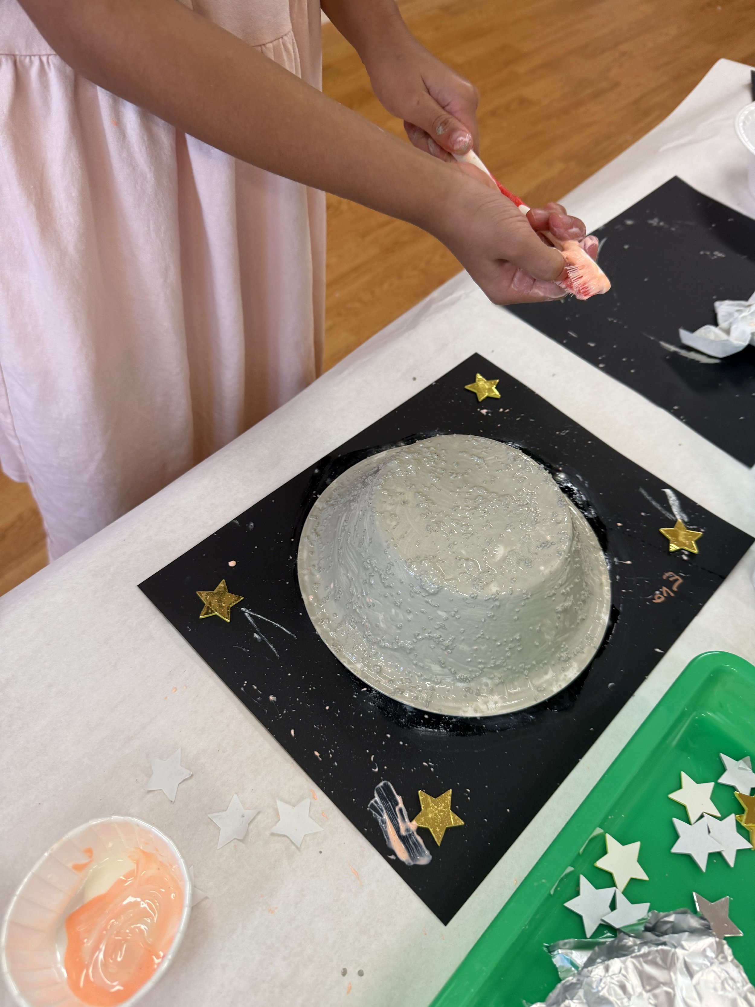 Child wearing a pink dress frosting a white cake with a piping bag, decorated with gold star confetti on the table.