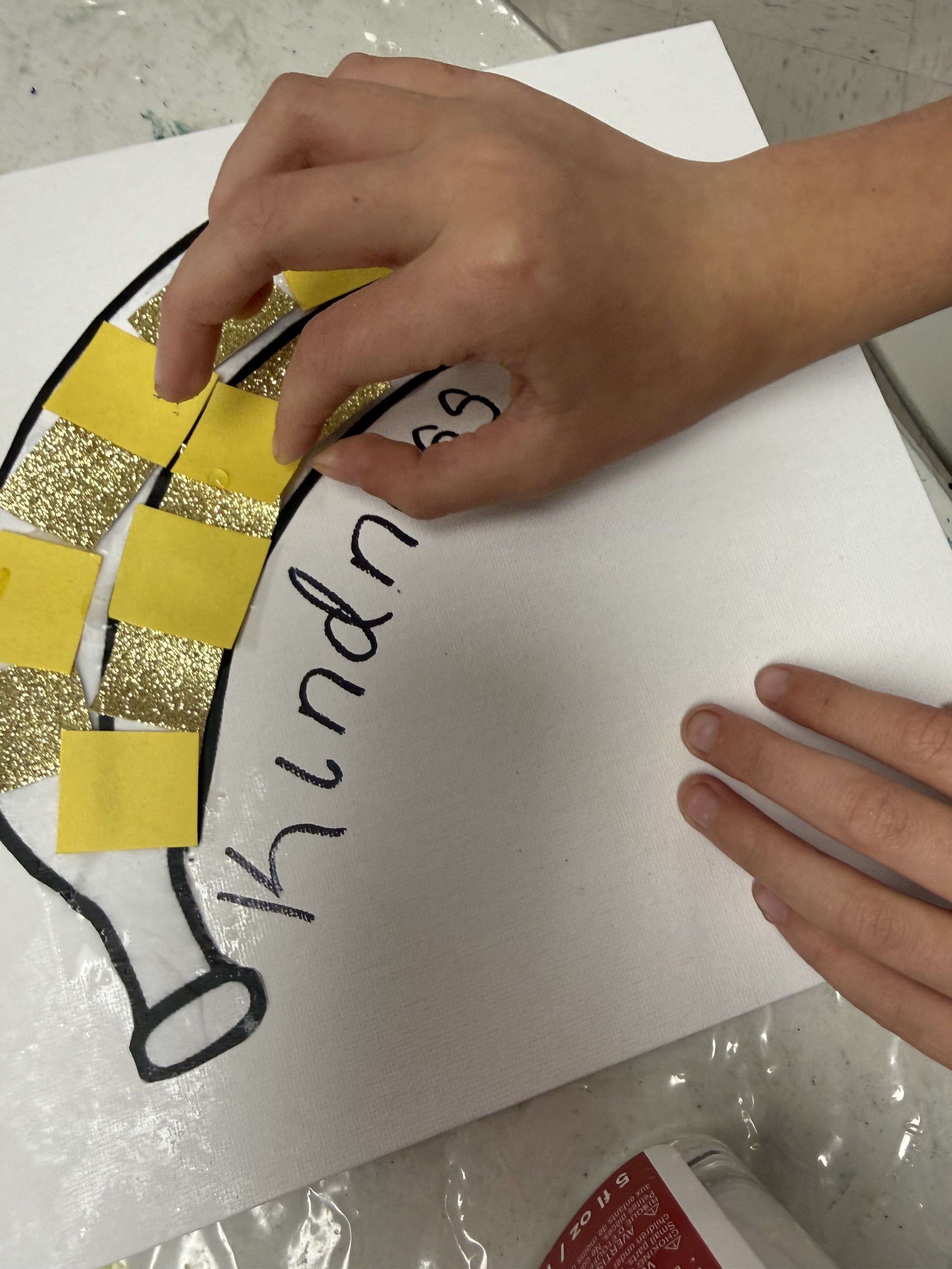 A person's hand placing yellow and gold glittery strips on a white sign that says 'Happy' with other words underneath, on a white surface.