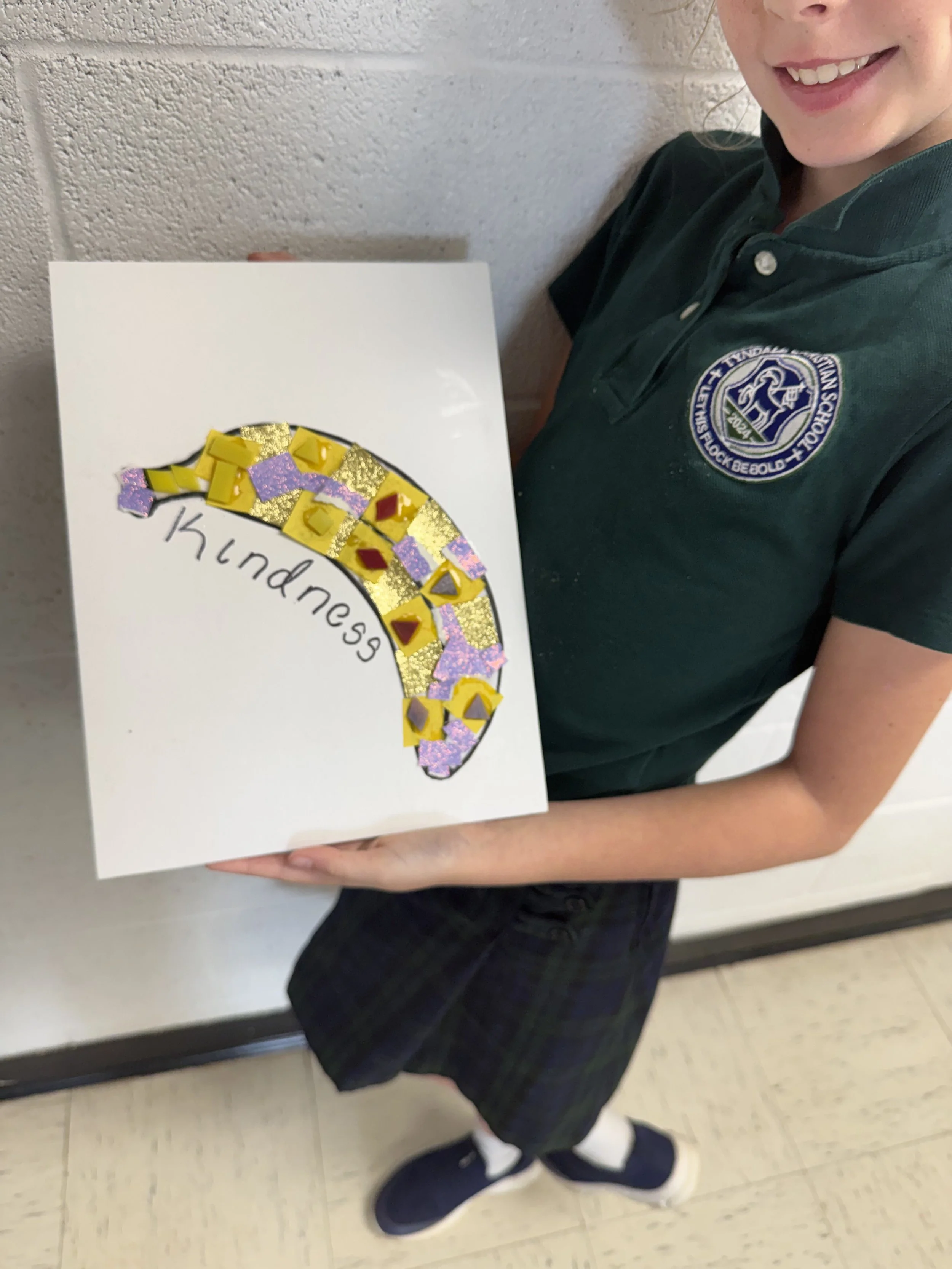 A girl in a school uniform holding a piece of artwork with a rainbow made of yellow, red, pink, and purple materials, with the word 'kindness' written underneath.
