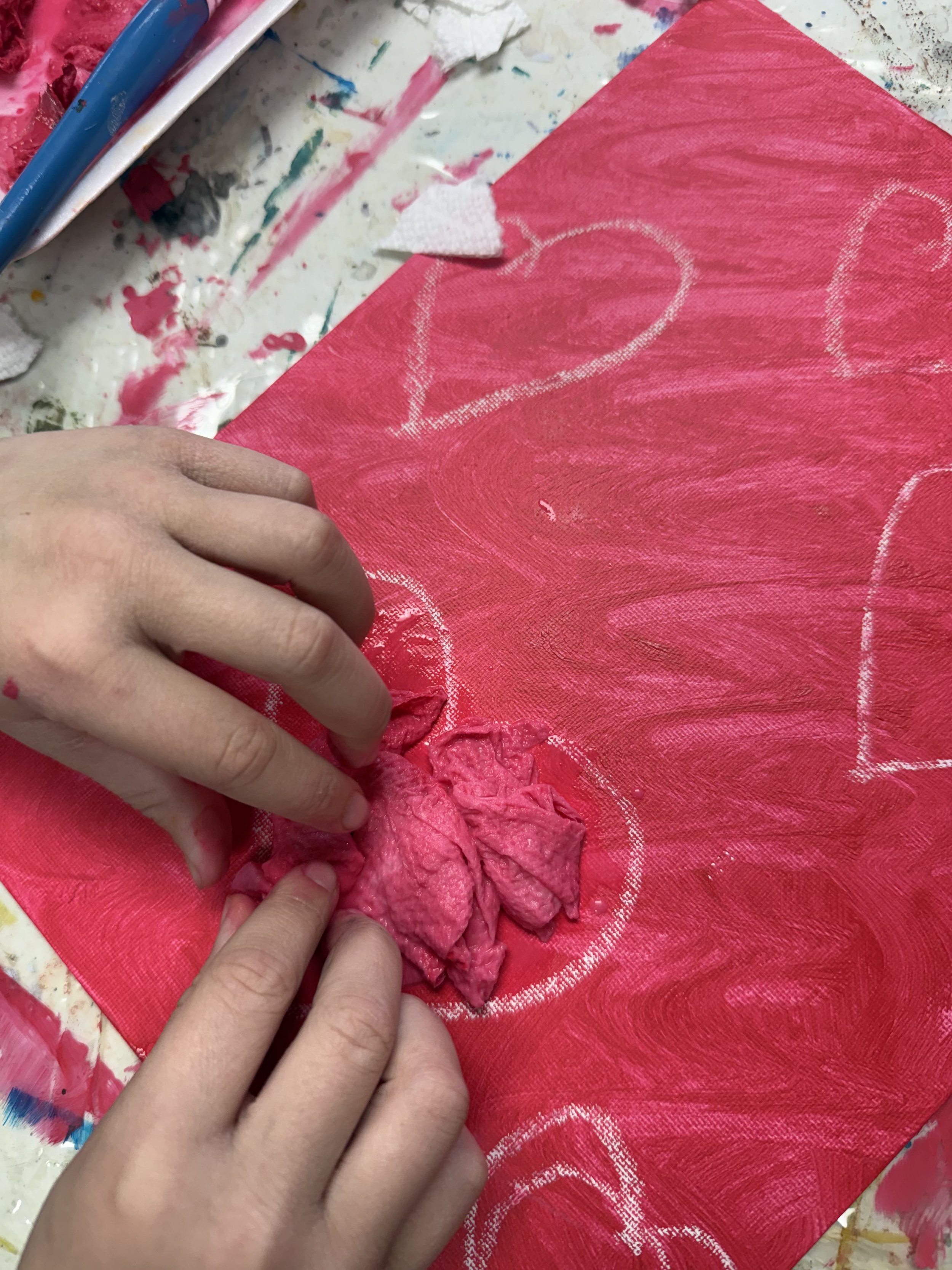 A person creating a heart design on a red fabric using white chalk, with paint and art supplies scattered around.