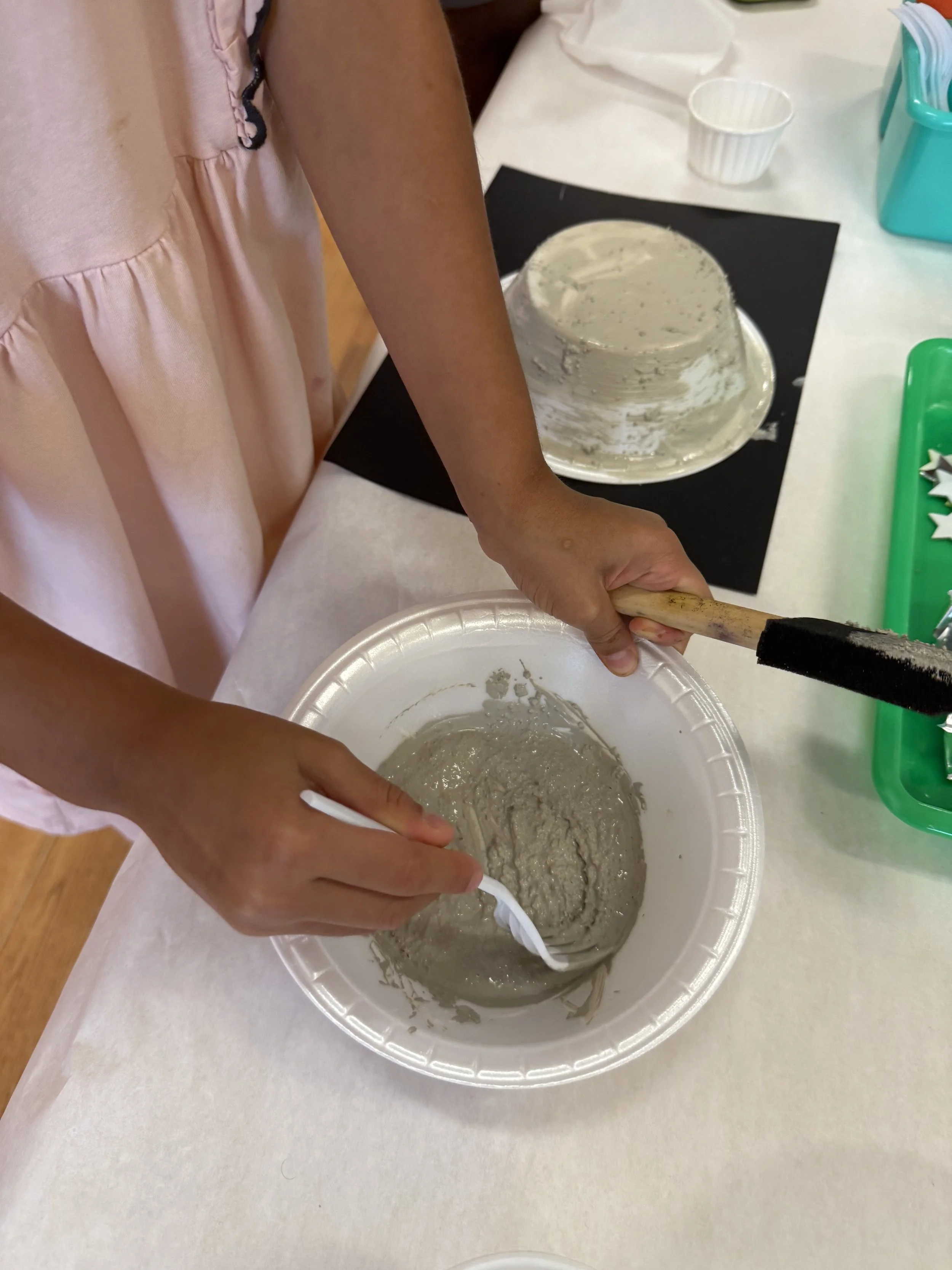 A person wearing a pink dress is stirring a grayish mixture in a white bowl with a white spoon. On the table, there is a cake with white frosting on a black board, a small white cup, and green containers, one with Christmas tree-shaped cutouts.