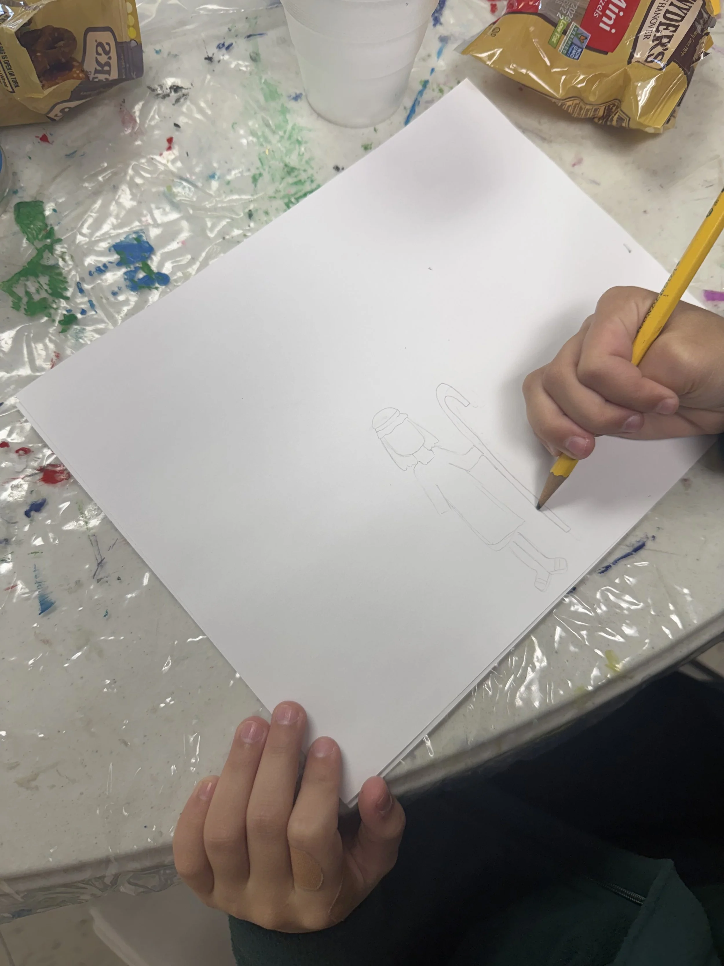 A person is drawing a person holding a cane on a white sheet of paper. The table is covered with a plastic tablecloth with paint stains, and there are snacks, a clear cup, and a bag of Goldfish crackers nearby.