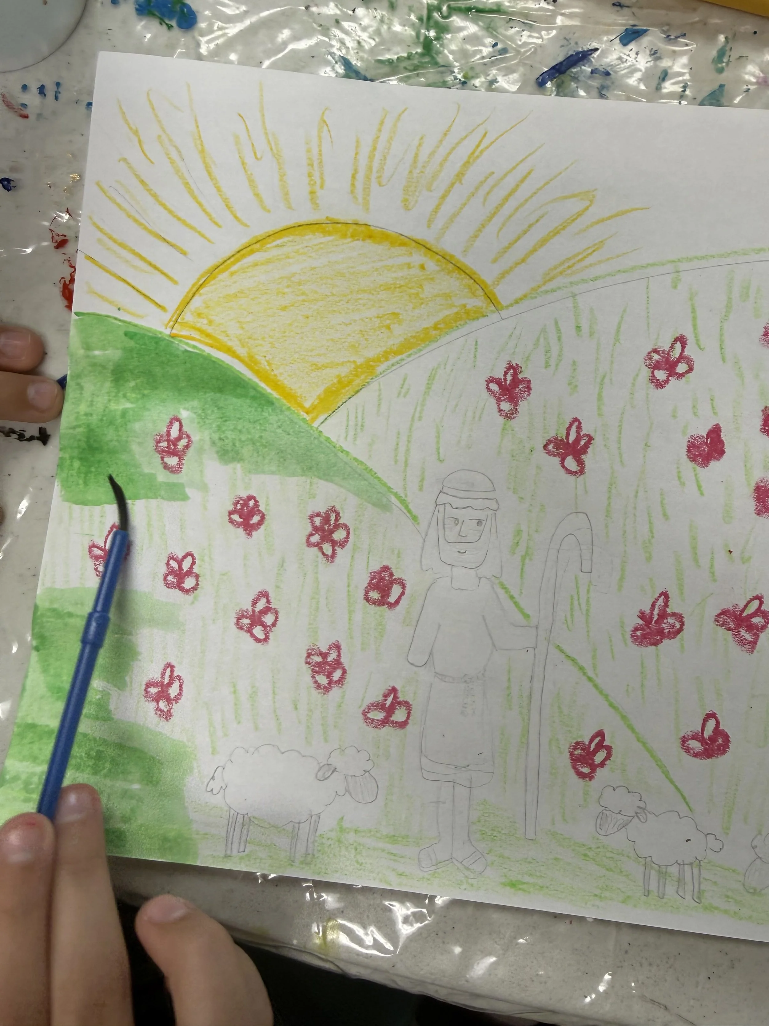 Child's drawing of a sunrise over green hills, with pink flowers, sheep, and a smiling person with a shepherd's staff.