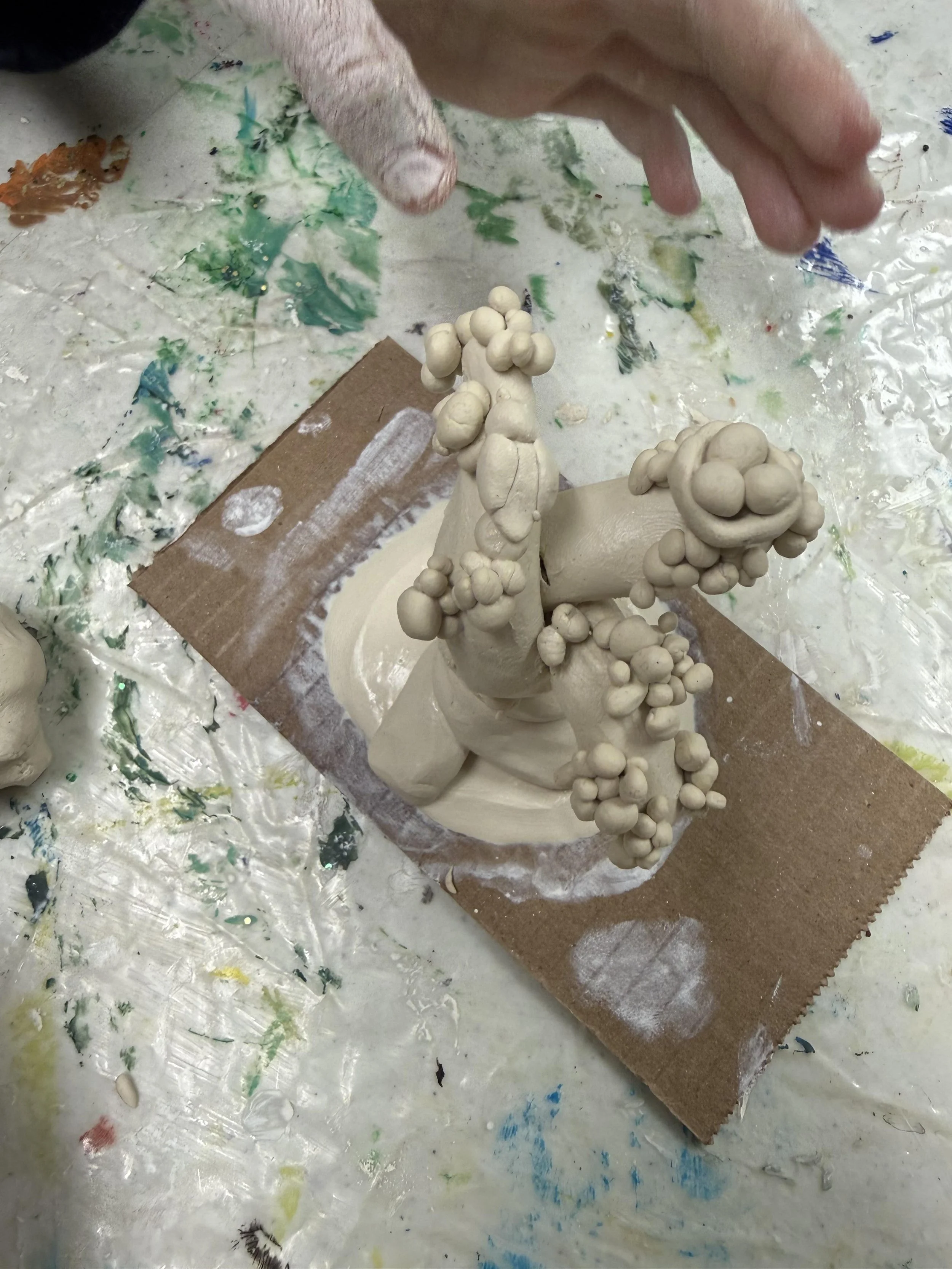 A ceramic sculpture in progress with multiple small clay spheres attached to a central structure, placed on a work surface with colorful paint splatters.