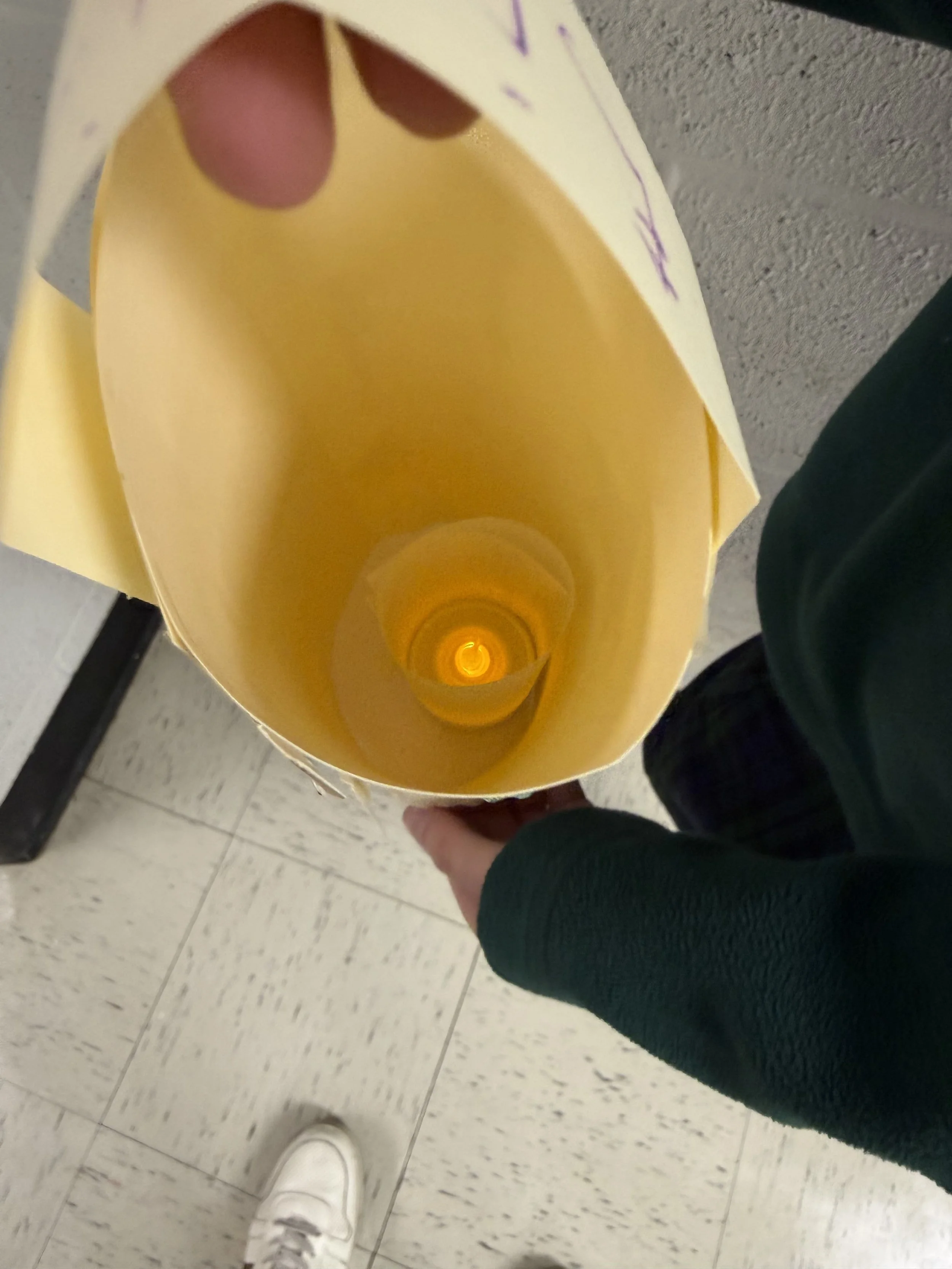 Person holding an empty yellow paper bag, looking down into it, with a tiled floor and a person's white sneaker visible in the background.