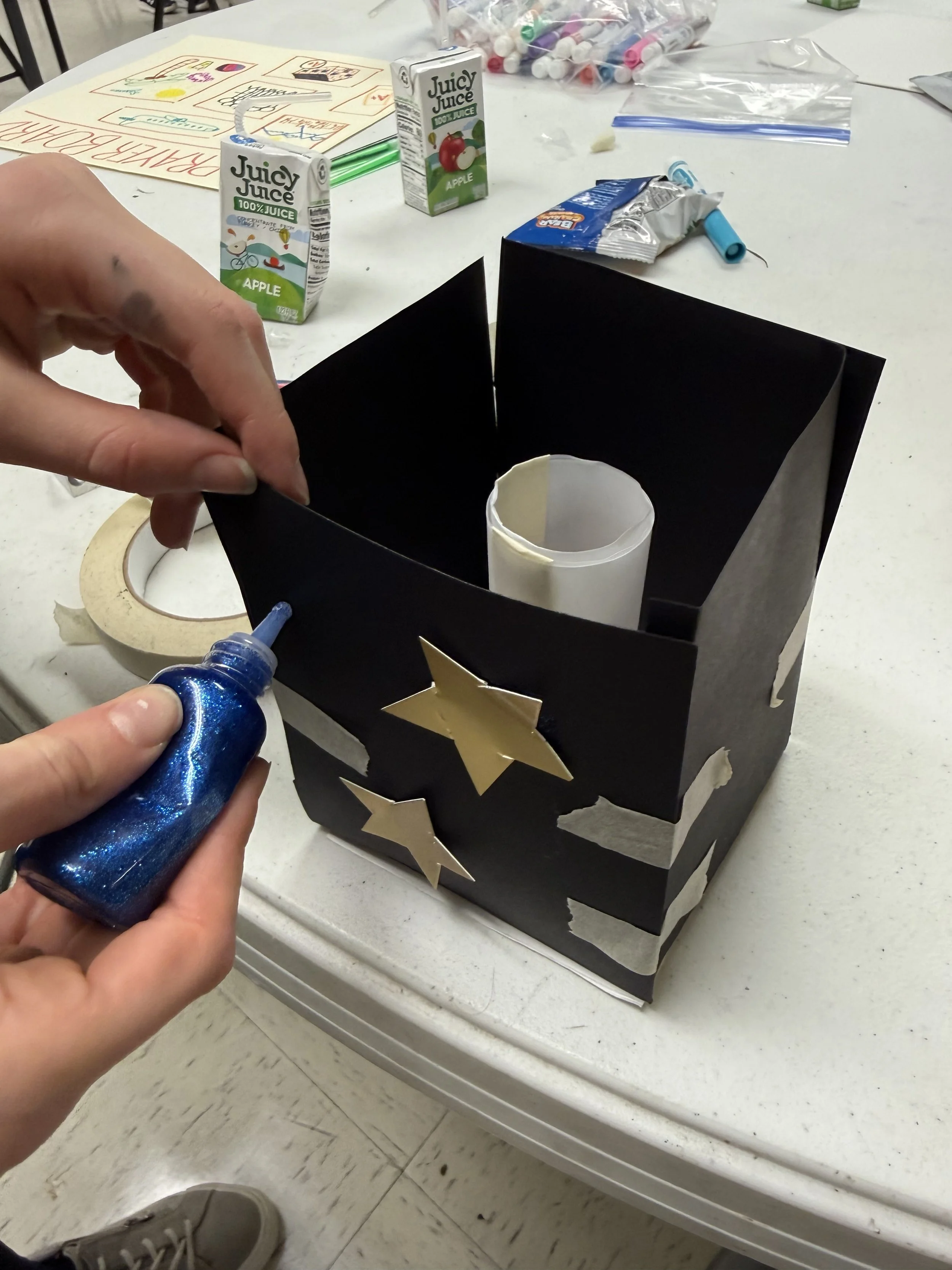 Person applying blue glitter glue to black craft cone decorated with gold star stickers, on a white table with craft supplies and juice boxes in the background.