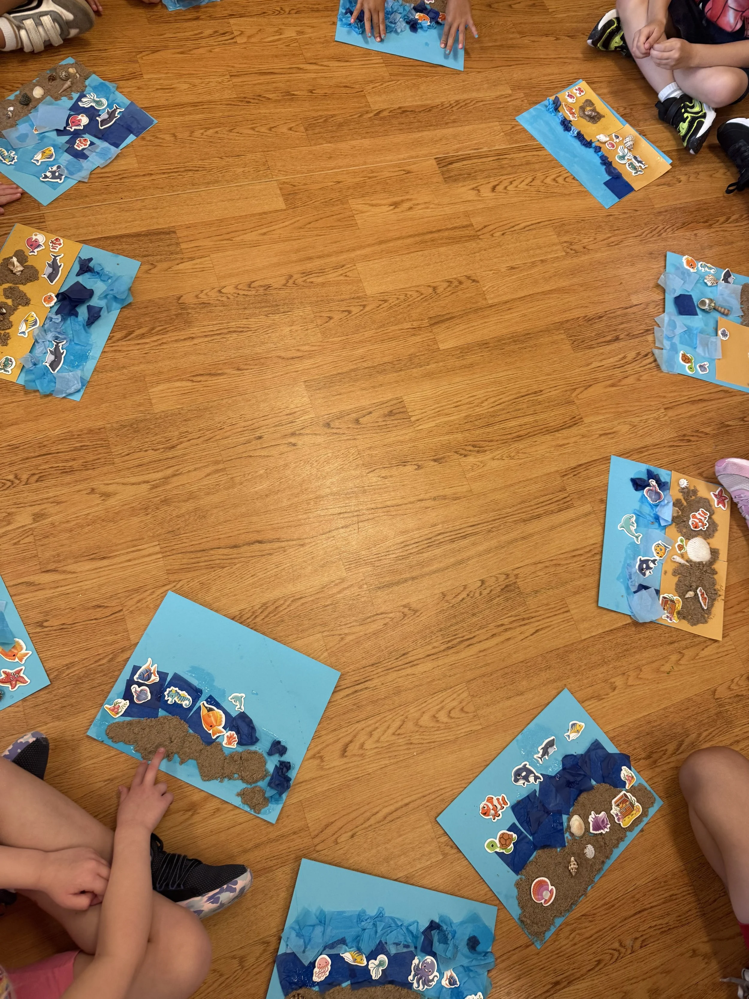 Children's craft activity with ocean scenes on blue and yellow paper, featuring stickers and tissue paper representing sea and ocean elements, arranged in a circle on a wooden floor.