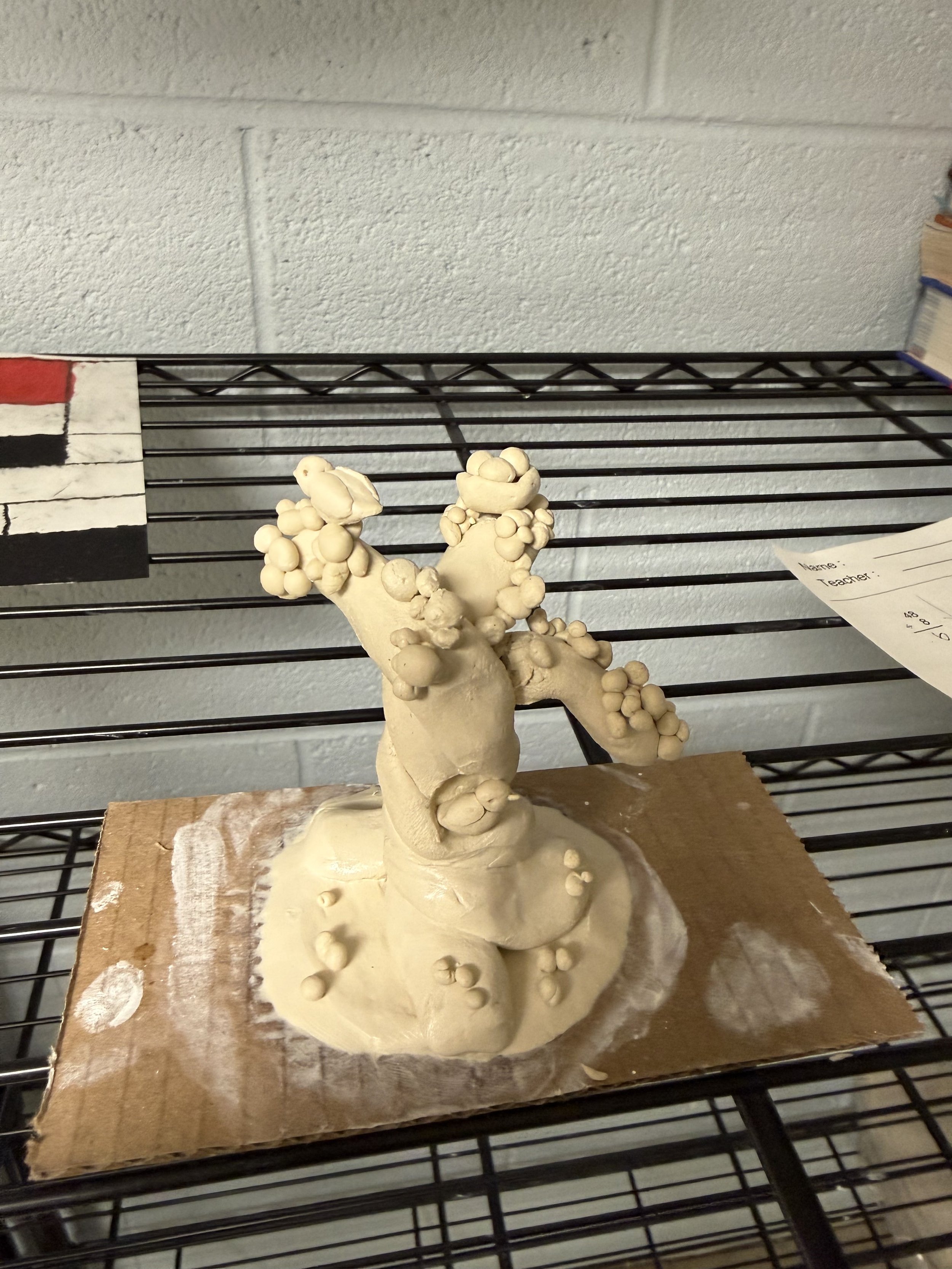 A clay sculpture of a tree with many spherical growths on its branches and trunk, placed on a cardboard base on a wire shelf.