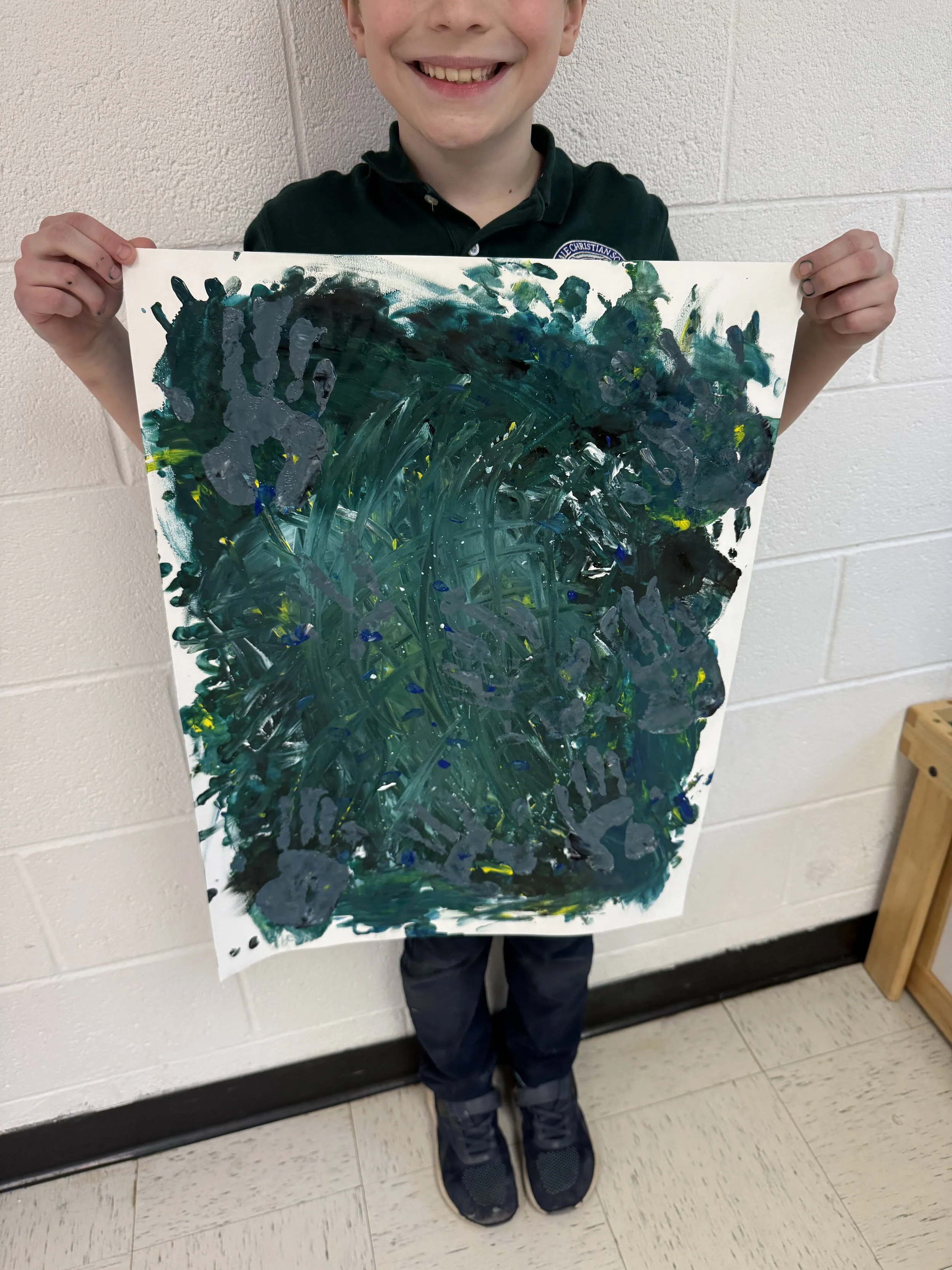 Young boy smiling and holding up a large abstract painting with dark green, gray, and small hints of yellow and blue colors.