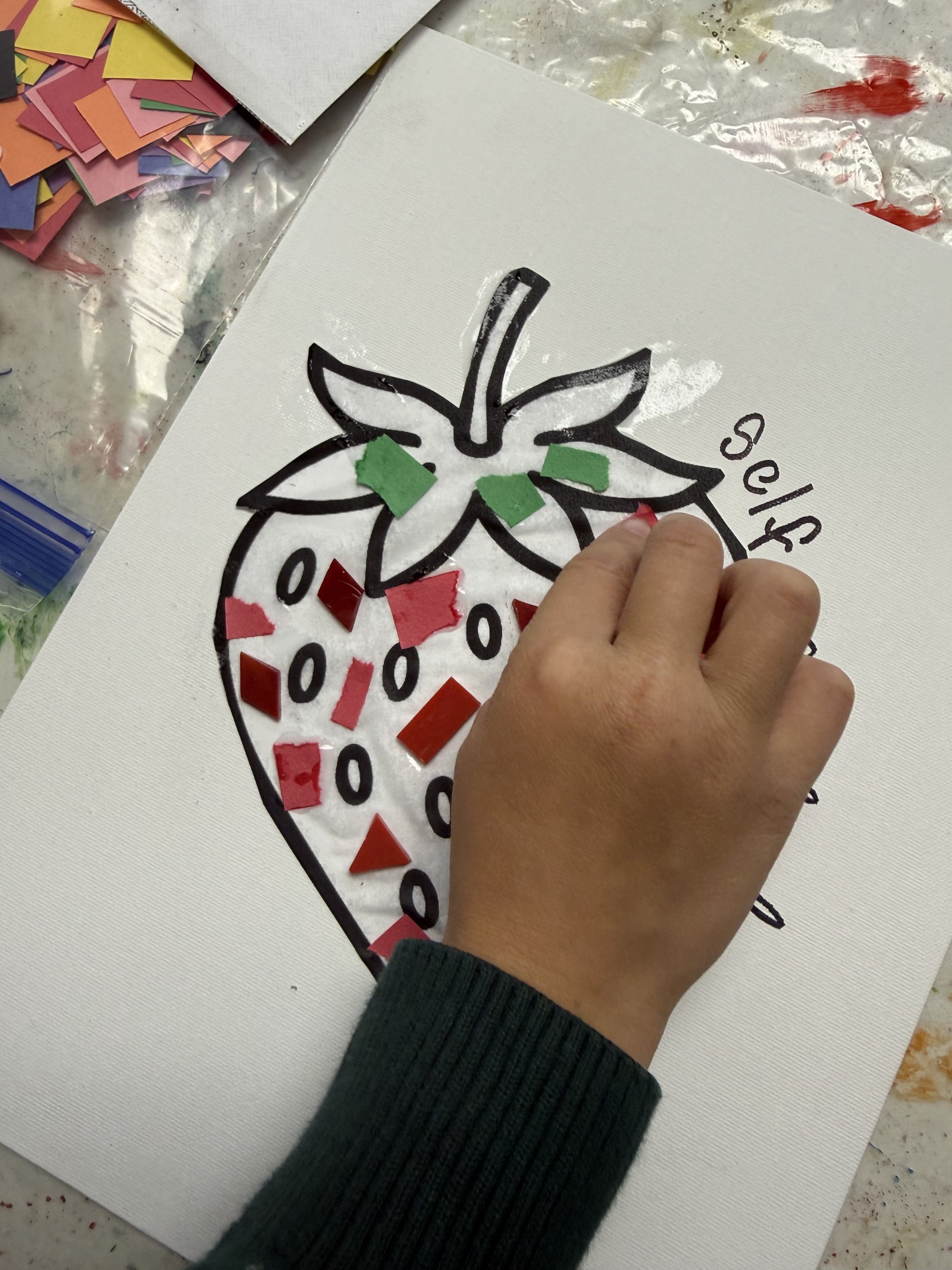 Child's hand gluing red and green mosaic pieces onto a strawberry outline on a white canvas.