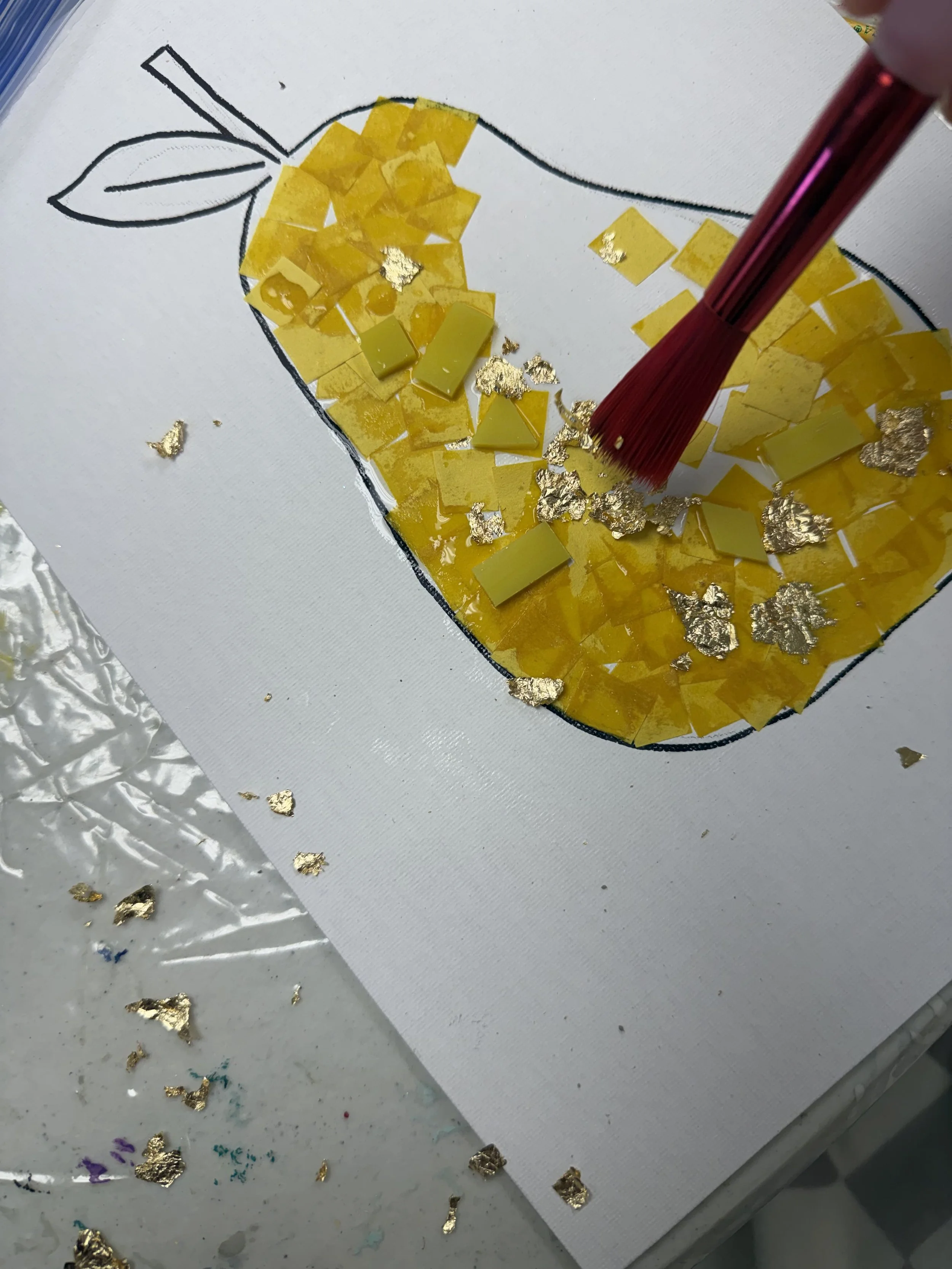 A collage art project of a yellow and gold apple being created with small yellow paper squares and gold foil pieces, with a red paintbrush applying adhesive on a white background.