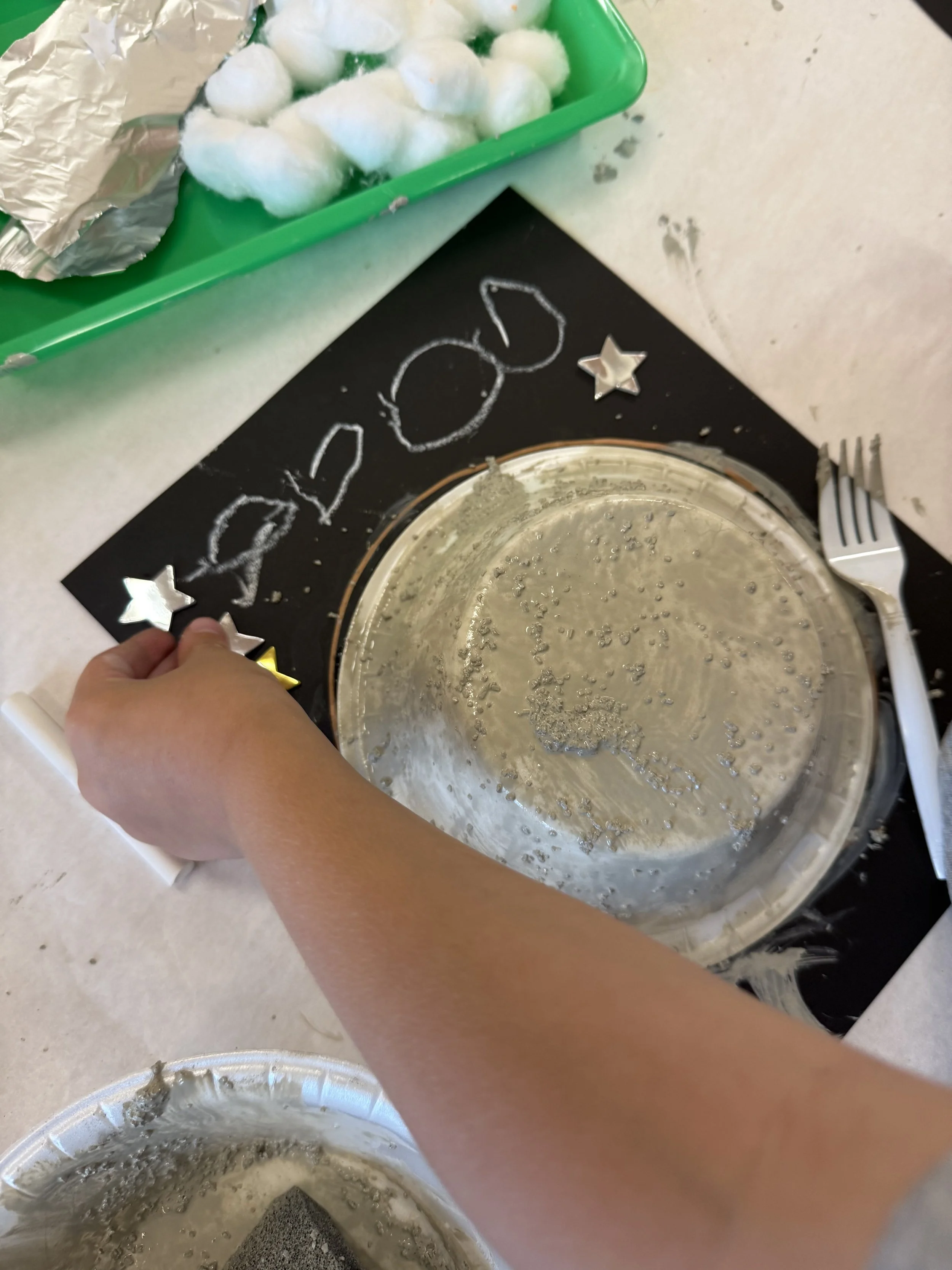 Empty pie pan on a black mat with the word 'FOOD' written in chalk, surrounded by small star decorations, with a fork nearby. In the background, there is a green tray with cotton balls and aluminum foil.