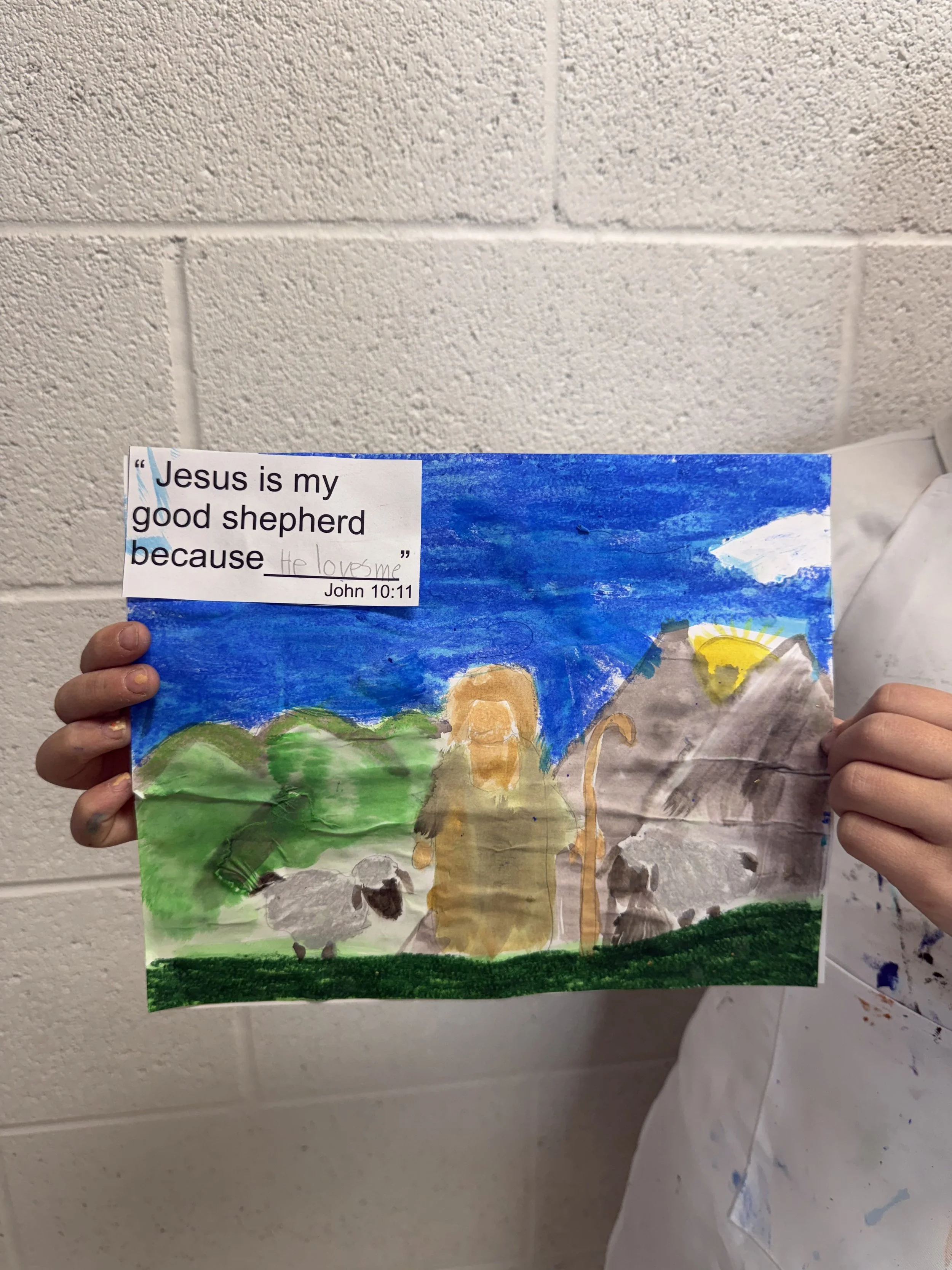A child's watercolor painting of Jesus with sheep in a green field, blue sky, mountains, and a sun, with a printed Bible quote from John 10:11 about Jesus being the good shepherd.