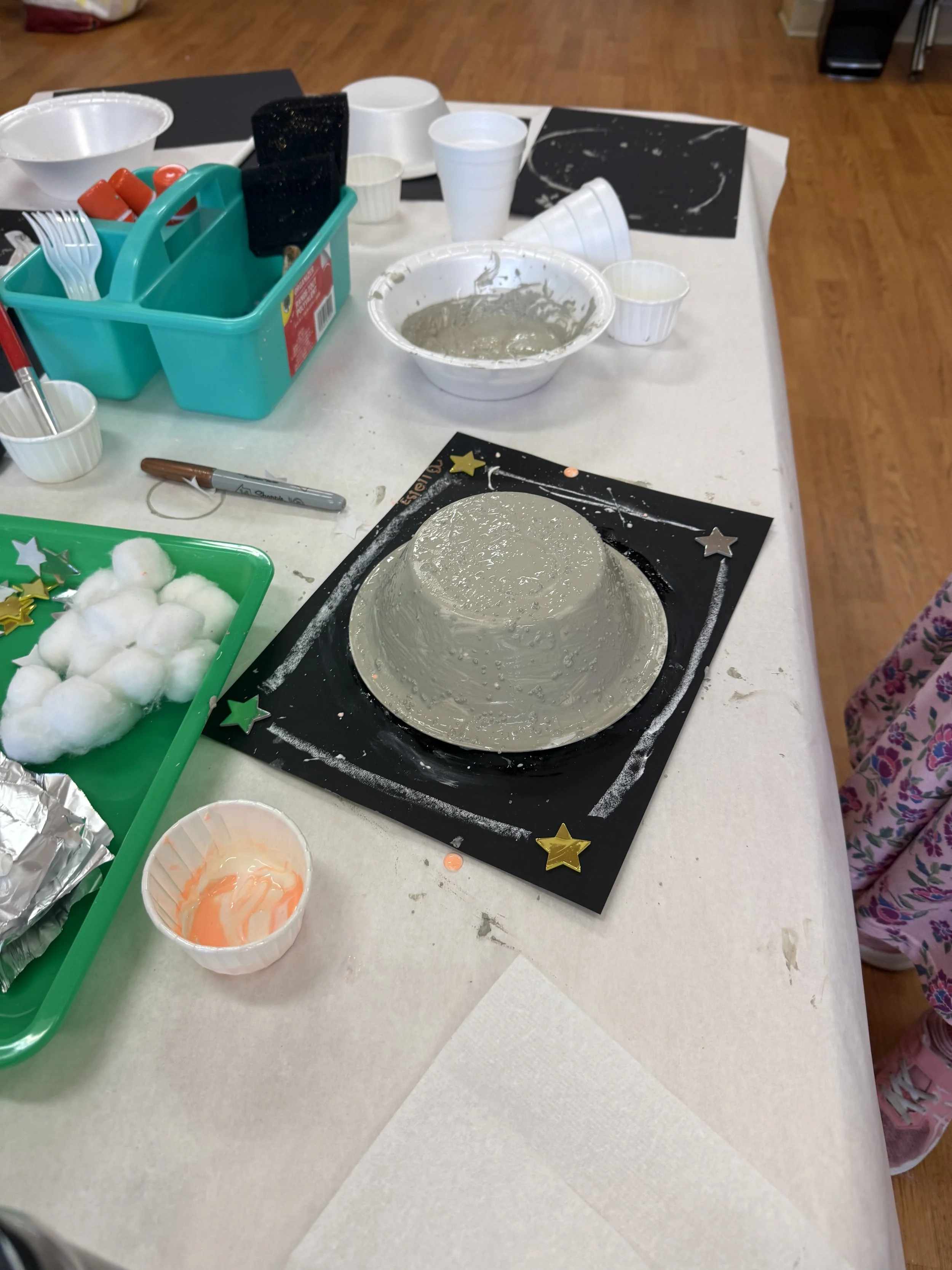 A tabletop with a black construction paper and a large upside down bowl covered in gray cement or plaster, decorated with star stickers, as part of a craft or project. Surrounding items include cotton balls, a marker, plastic cups, bowls, and various