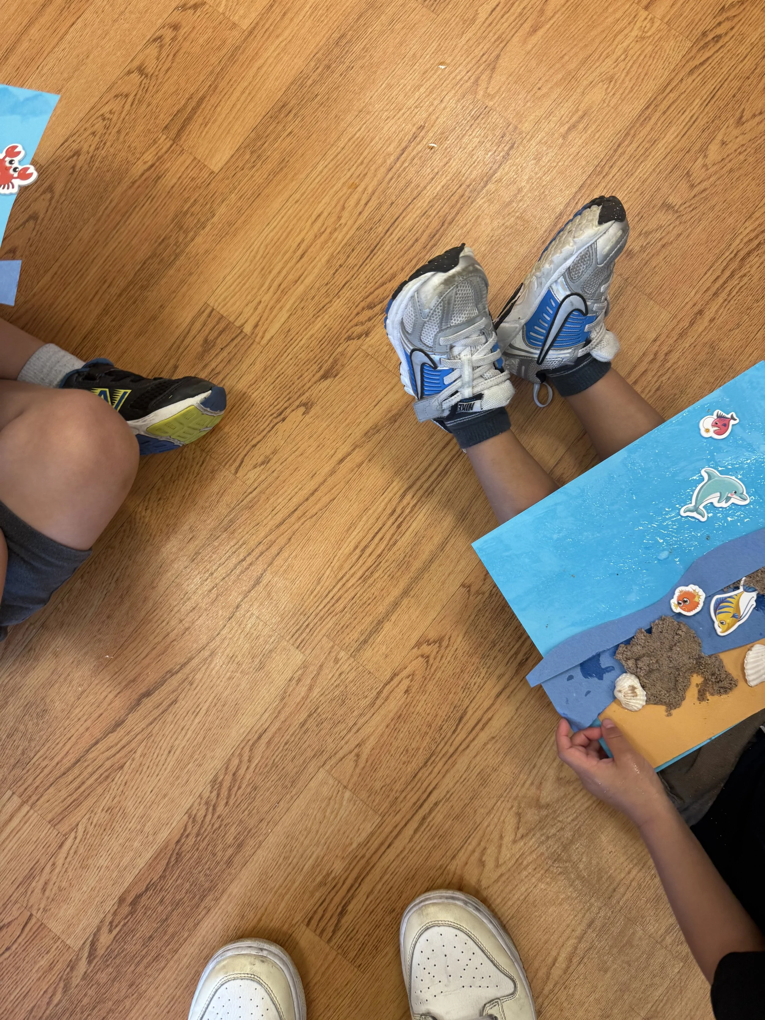 Children sitting on a wooden floor, with a blue display board decorated with fish and shark stickers, pieces of foam, shells, and sand.