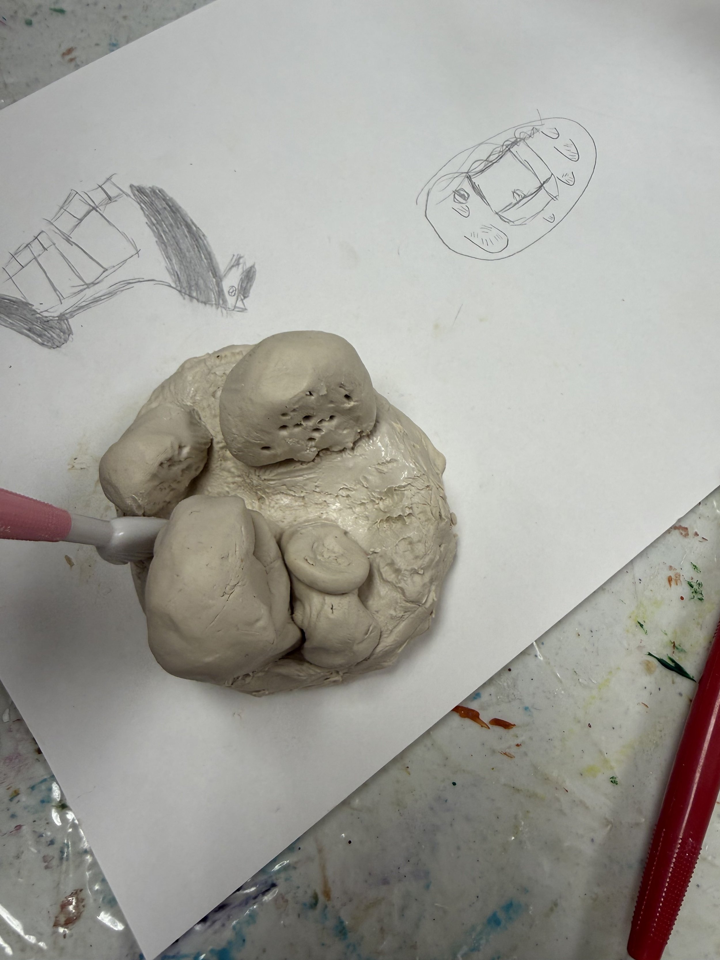 Clay sculpture in progress resembling a humanoid figure with rough features, placed on white paper with two pencil sketches of different character faces nearby, on a messy work surface.