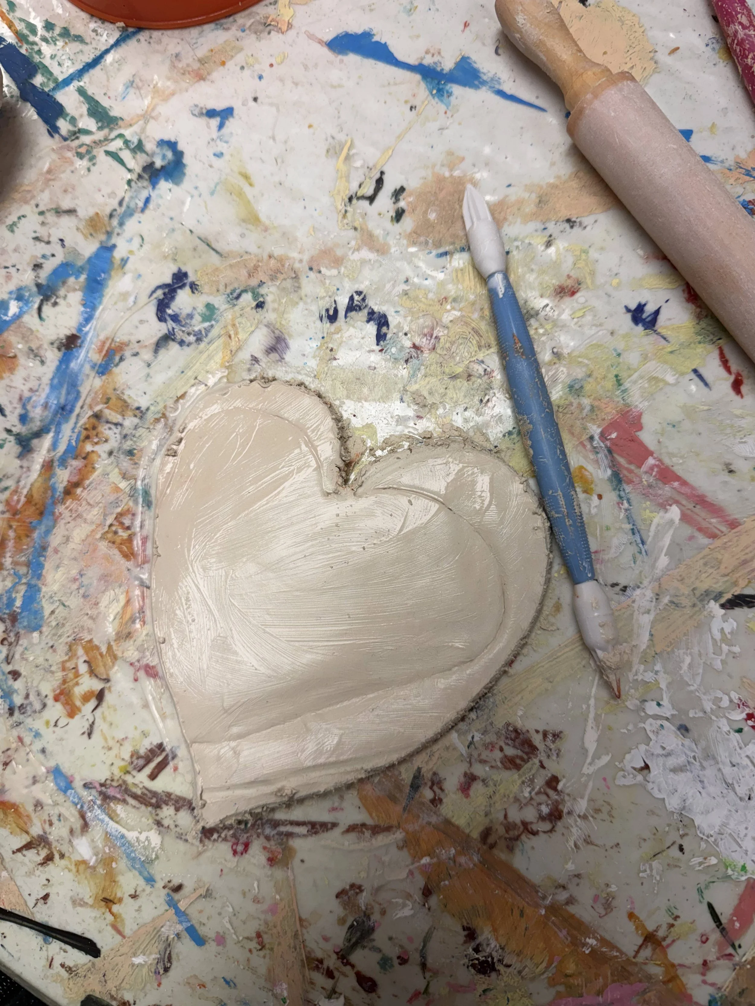 Clay heart sculpture on a cluttered artist's workspace with paint and tools.