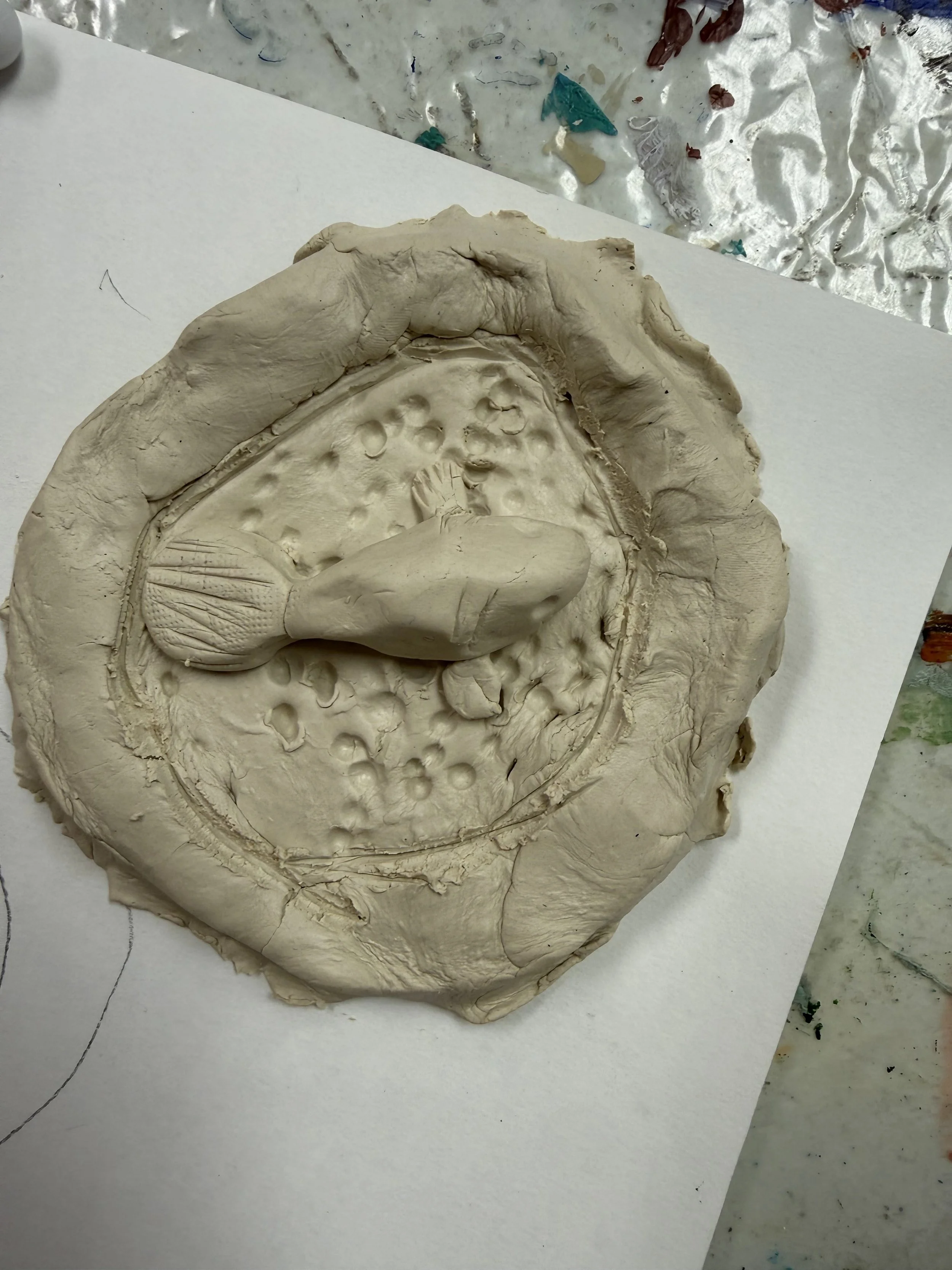 Unfinished ceramic sculpture of a fish, with a textured tail, on a round base with indented and dotted surface details.