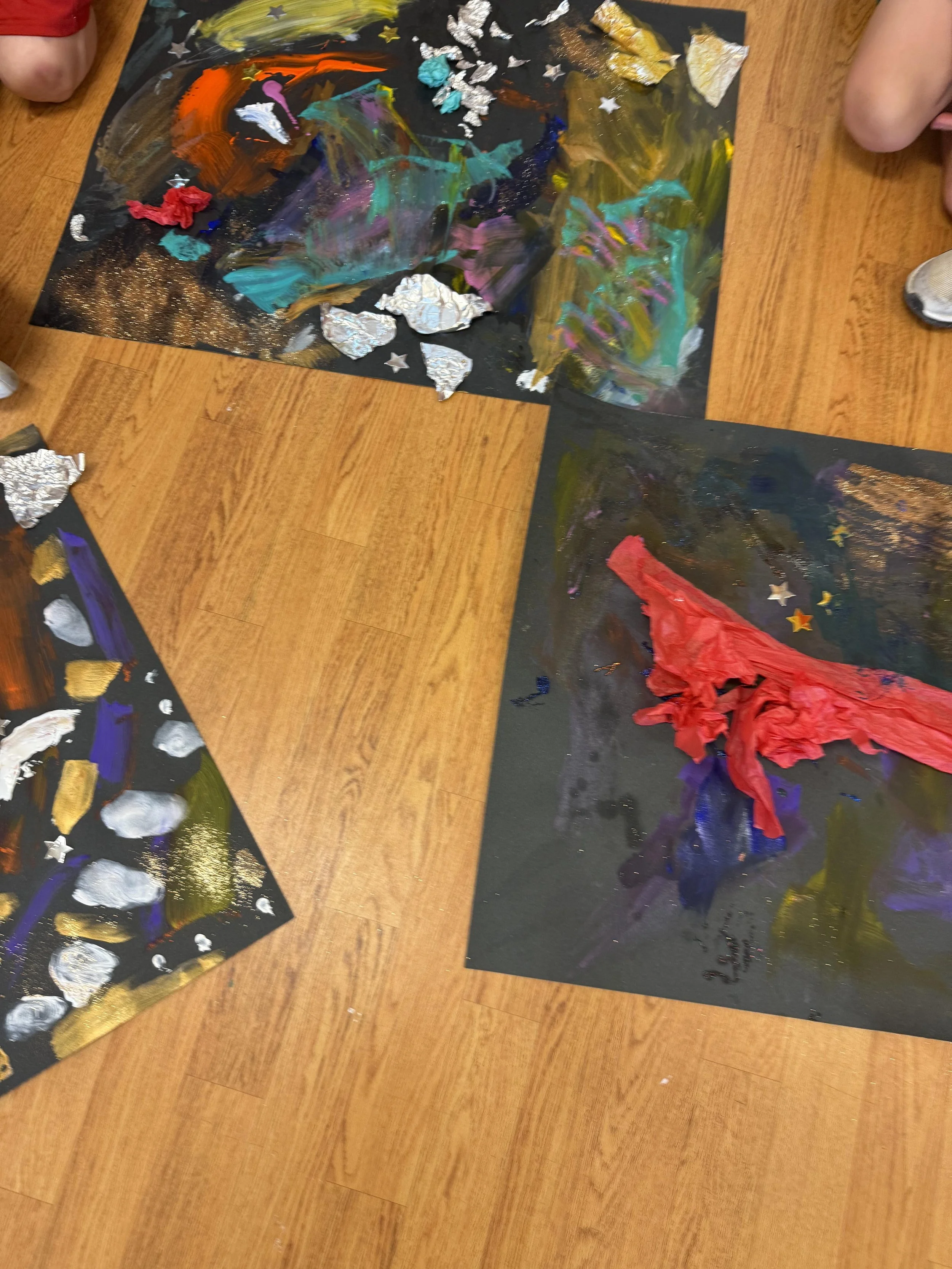 Three abstract paintings on black paper with paint, foil, and tissue paper, placed on a wooden floor.