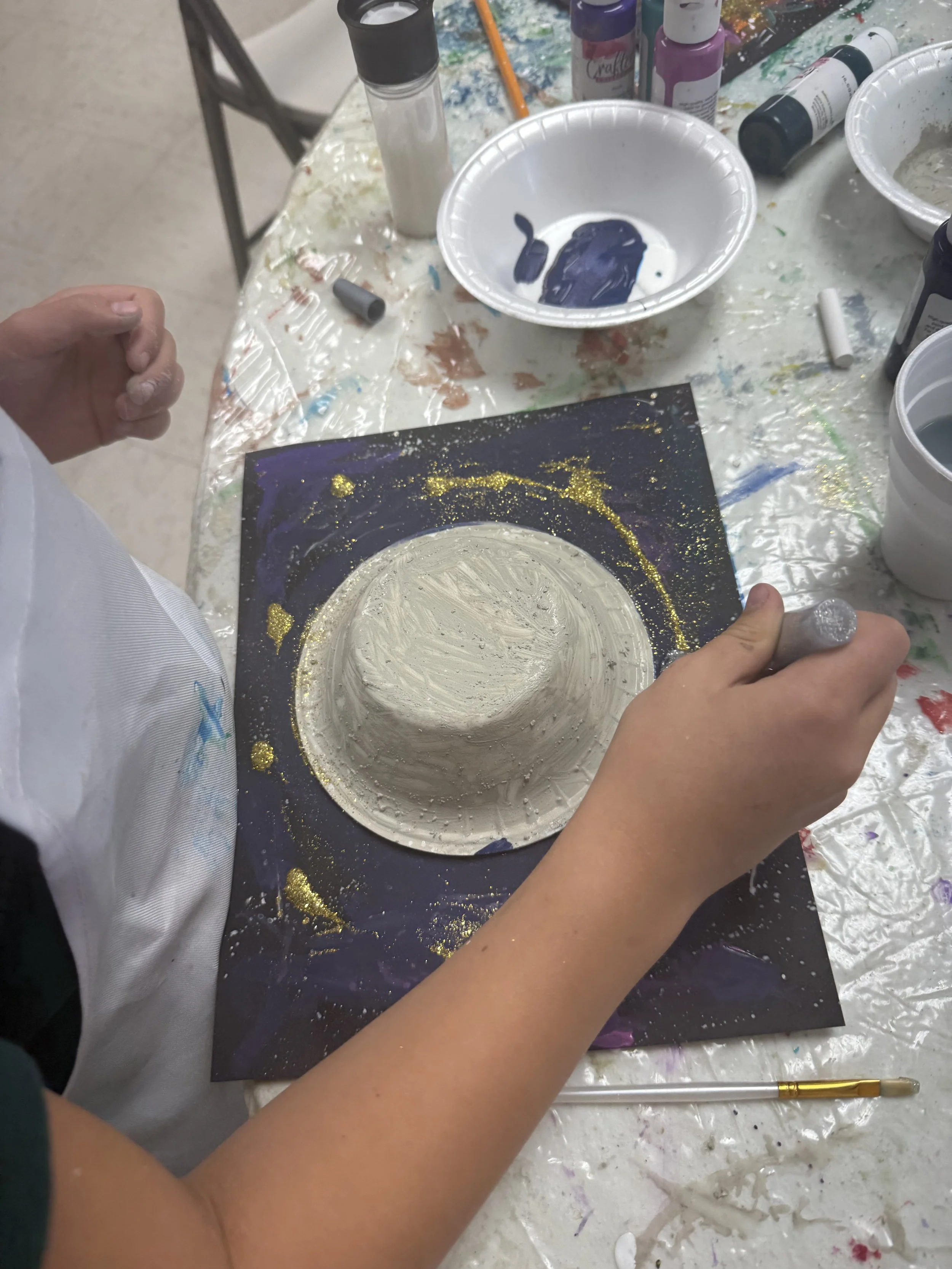 Child painting a circular object with white paint on a black canvas, surrounded by gold and purple paint splatters, with art supplies scattered on a table.