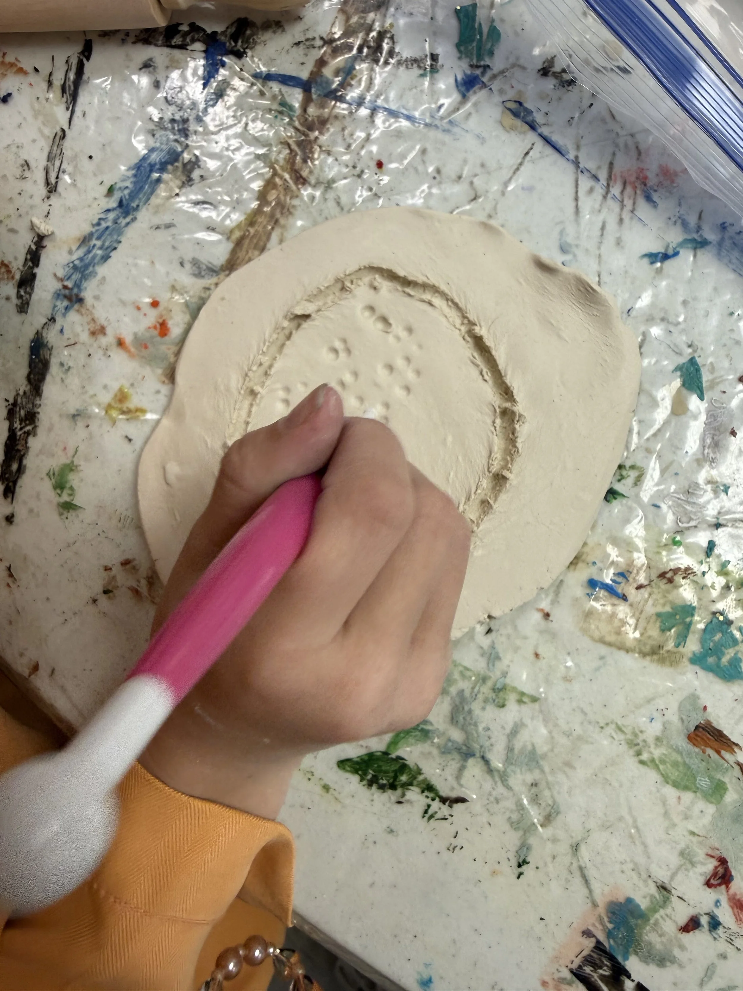 Person using a pink embossing tool to etch a heart shape with small dots into a sheet of white clay on a cluttered, paint-splattered work surface.