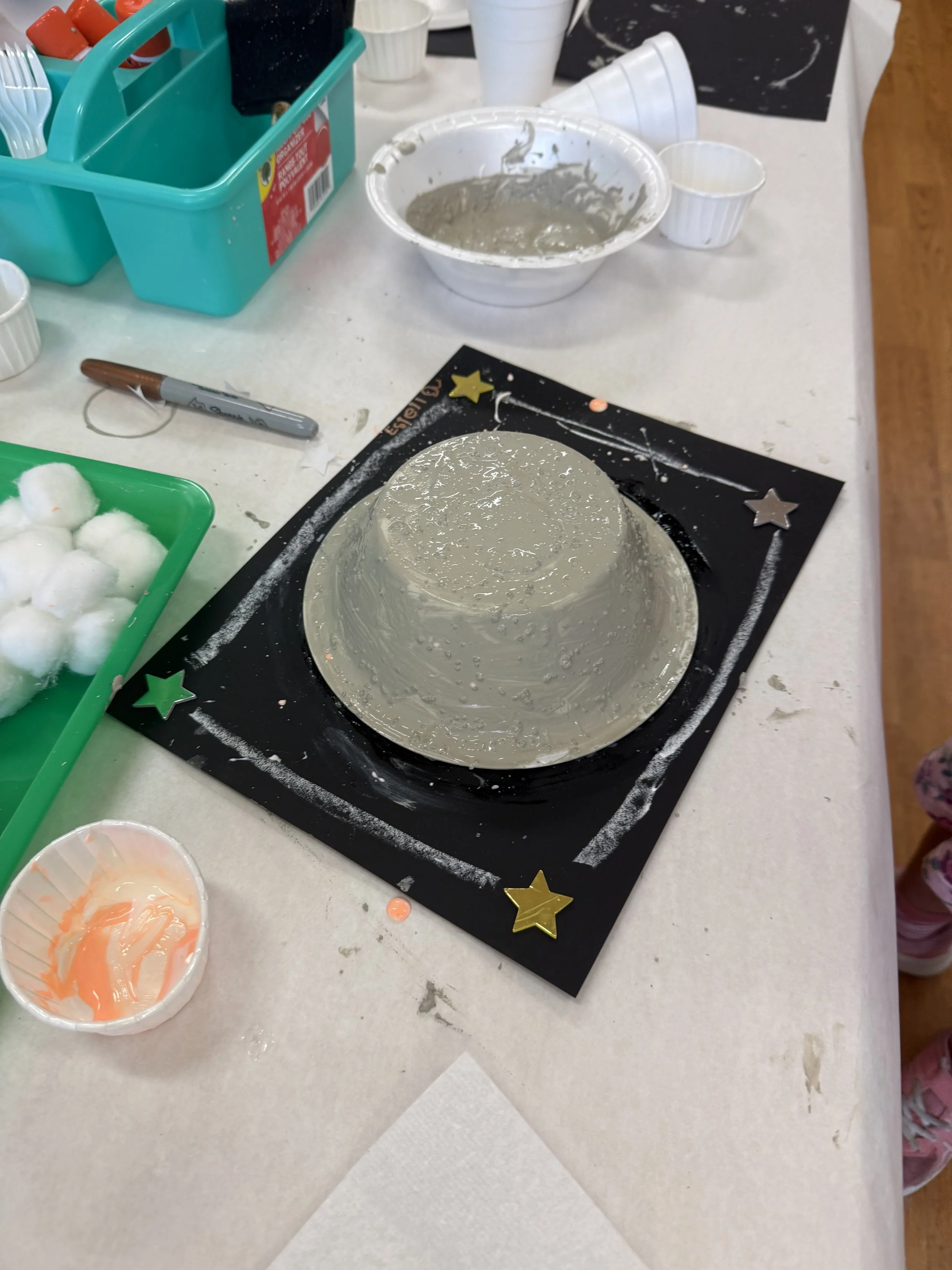 A decorated party hat mold sitting on a black tray with star decorations, surrounded by crafting supplies such as cotton balls, paint, brushes, and cups on a white table.