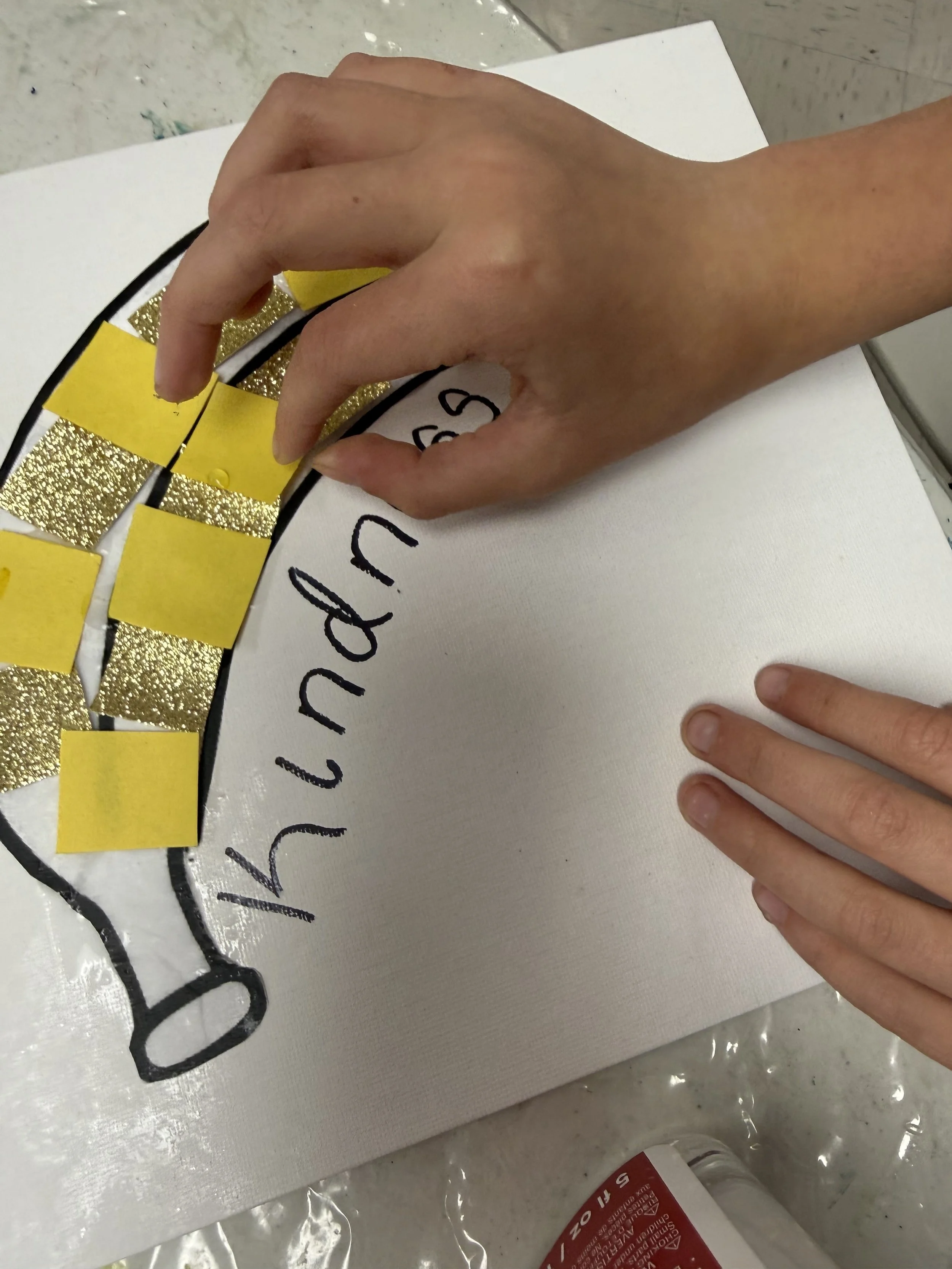 A person working on a craft project with a white canvas. Their left hand holds a black marker, writing or drawing on the canvas, which has the words 'Happy' and 'B' visible. Their right hand rests nearby. There are gold glittery and yellow paper piec