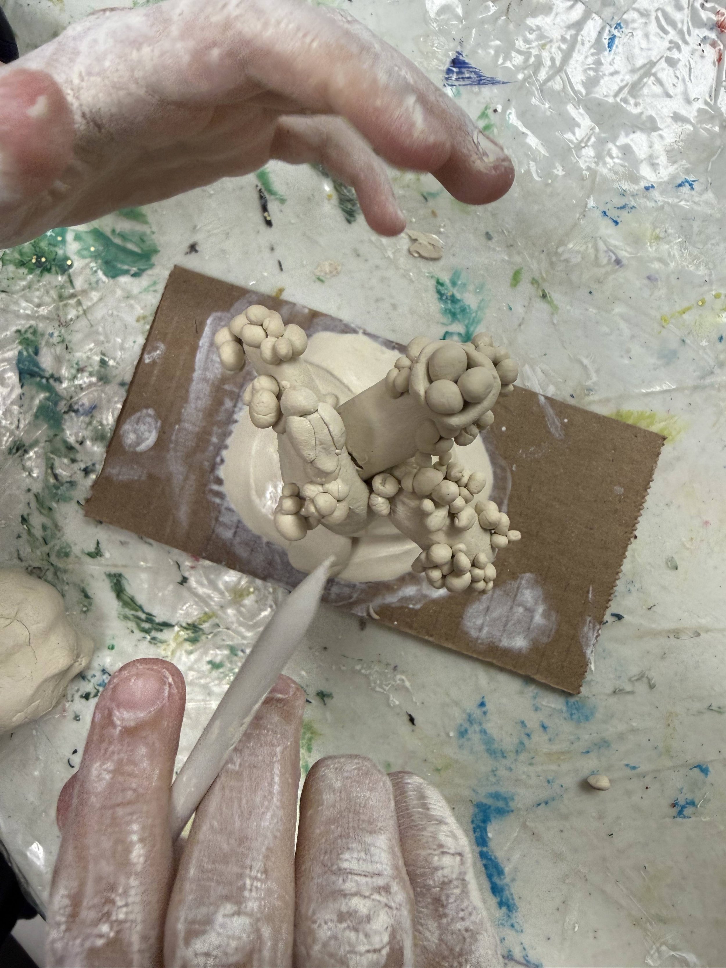 A person creating a ceramic sculpture with clay, using a tool to shape the piece on a work surface covered in clay and paint splatters.