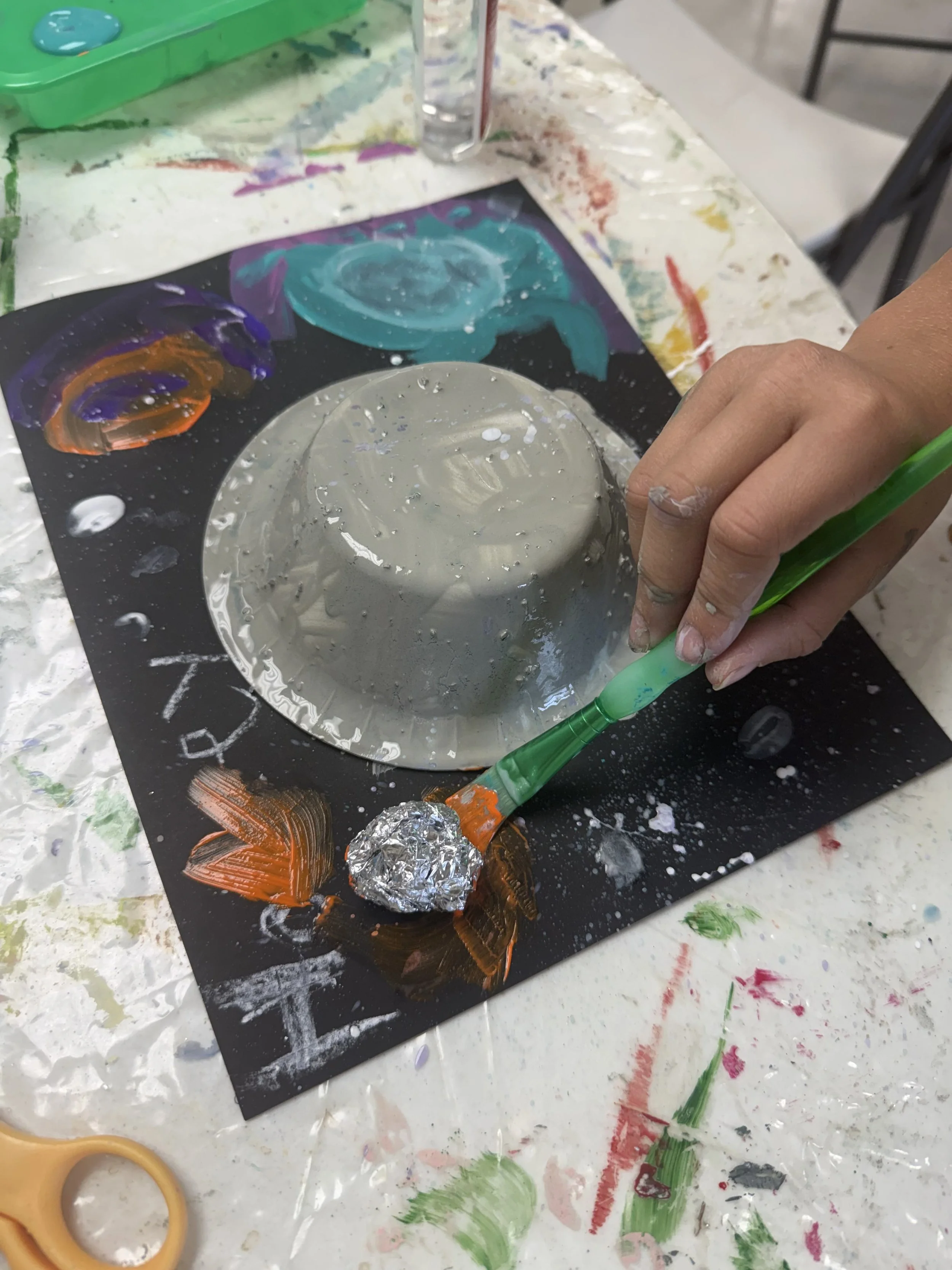 Child's hand painting a gray ceramic bowl with a green paintbrush on a black paper with colorful swirls and white writings, surrounded by art supplies like scissors, tape, and paint.