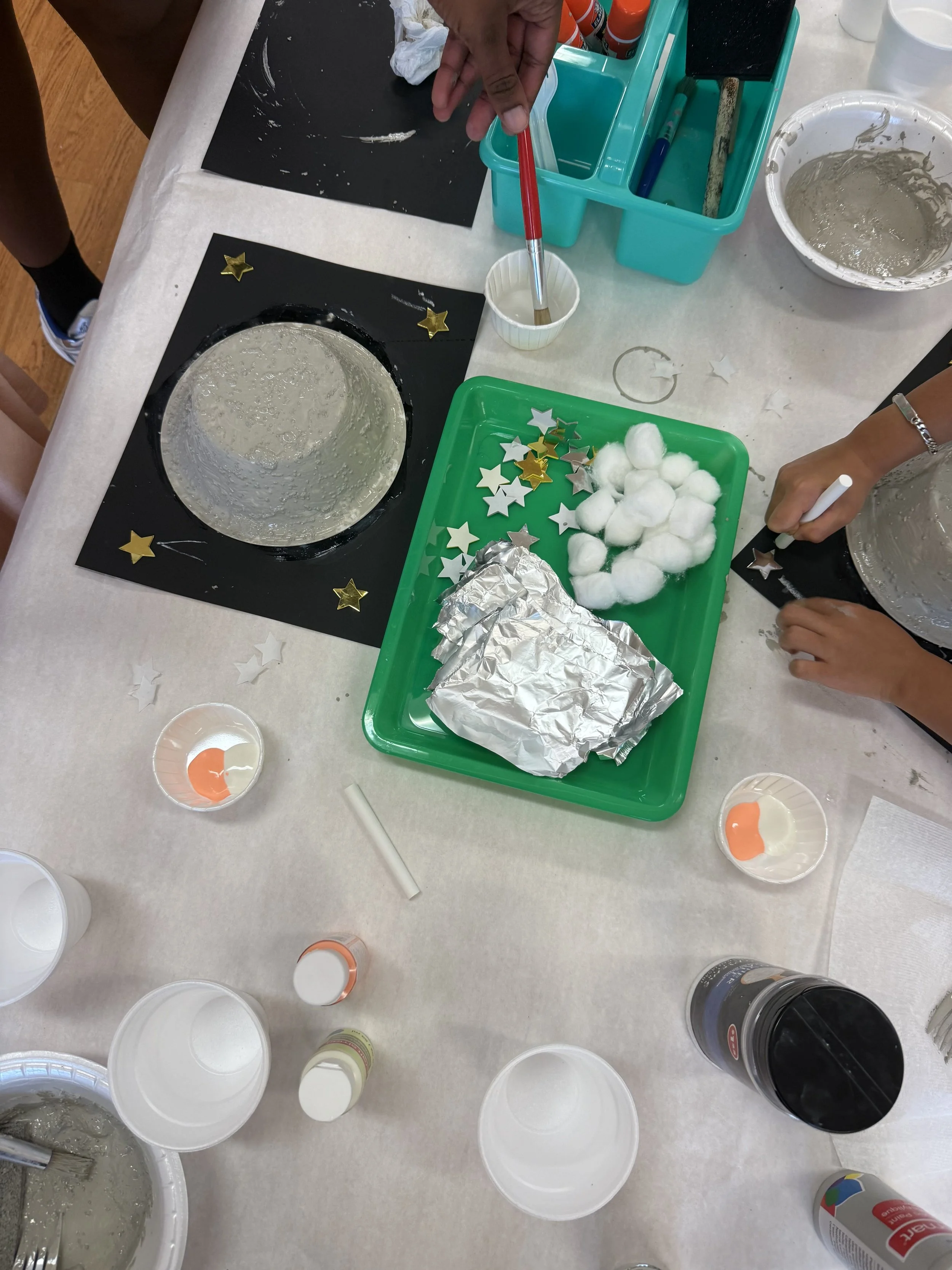 Children engaging in arts and crafts, decorating black paper with star-shaped confetti, cotton balls, and other supplies on a table.
