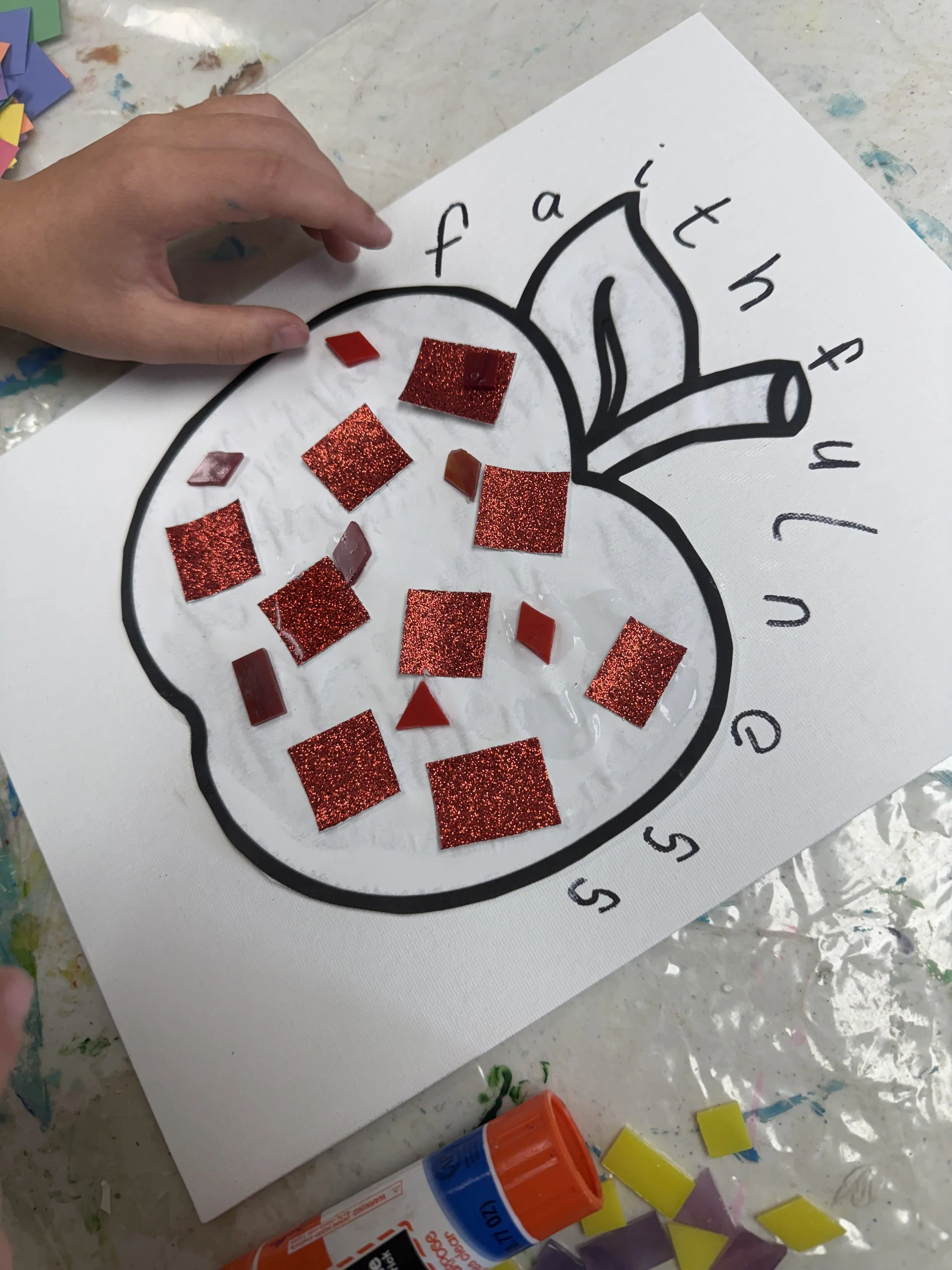 A hand glues red glittery and solid red paper squares onto a comic-style drawing of a cherry with the text 'f a i t h' surrounding it, on white paper.