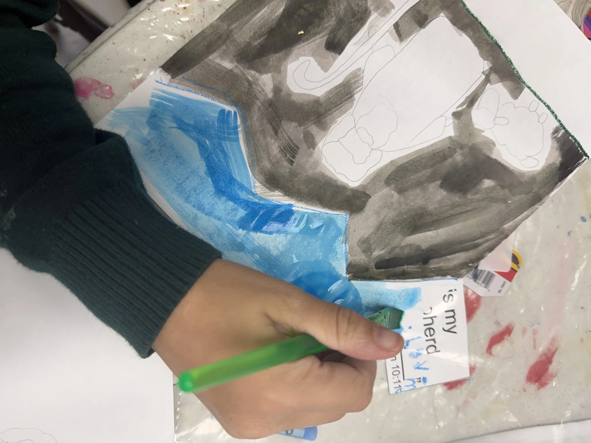 Close-up of a child's hand holding a green marker, coloring on a paper bag with a partly painted blue surface and a landscape outline, next to a partially visible black sweater sleeve. The paper bag shows some text and is placed on a plastic-covered 