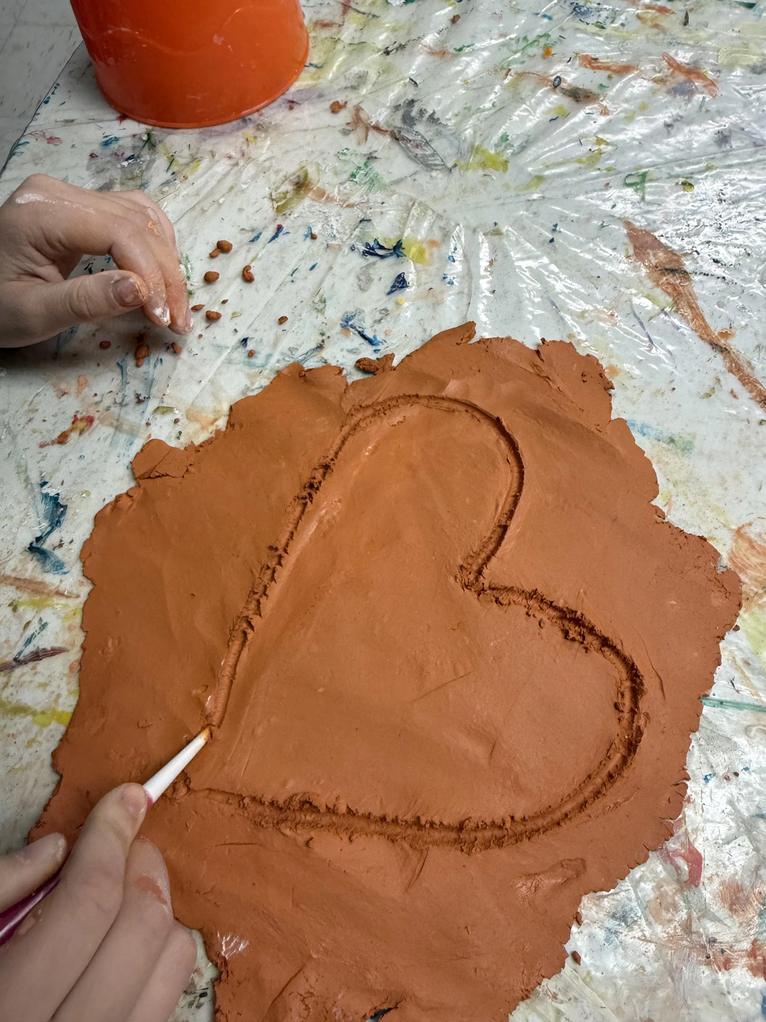 A person is tracing a heart shape in wet clay on a table covered with a colorful plastic sheet, with their left hand visible around the top left corner.