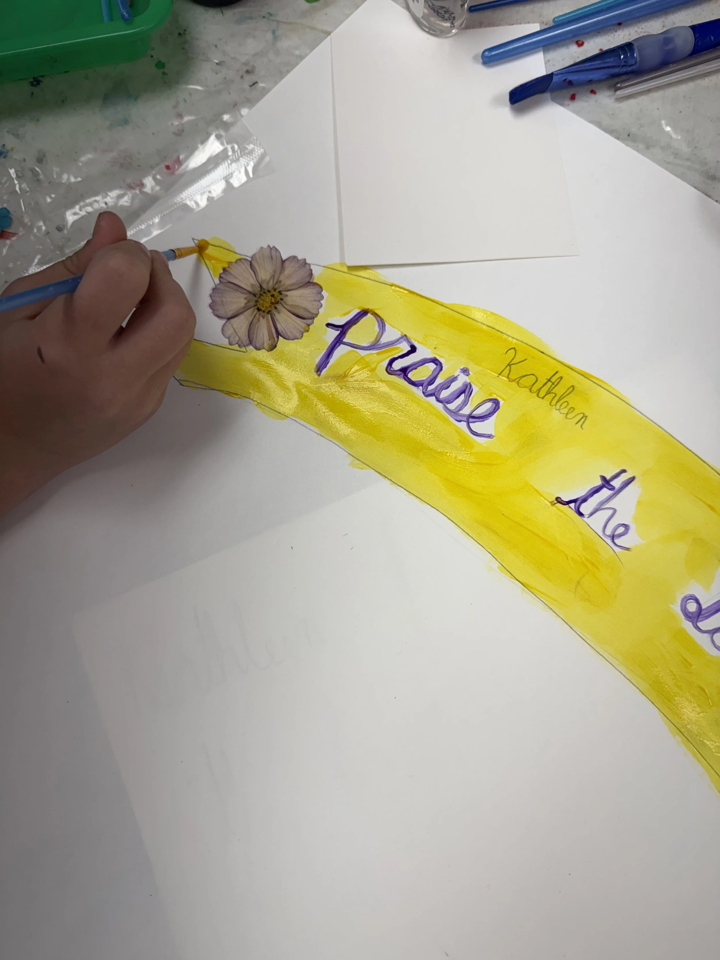 A person is painting a handmade card with yellow watercolor, writing 'Please' in purple with a flower graphic on a white sheet of paper.