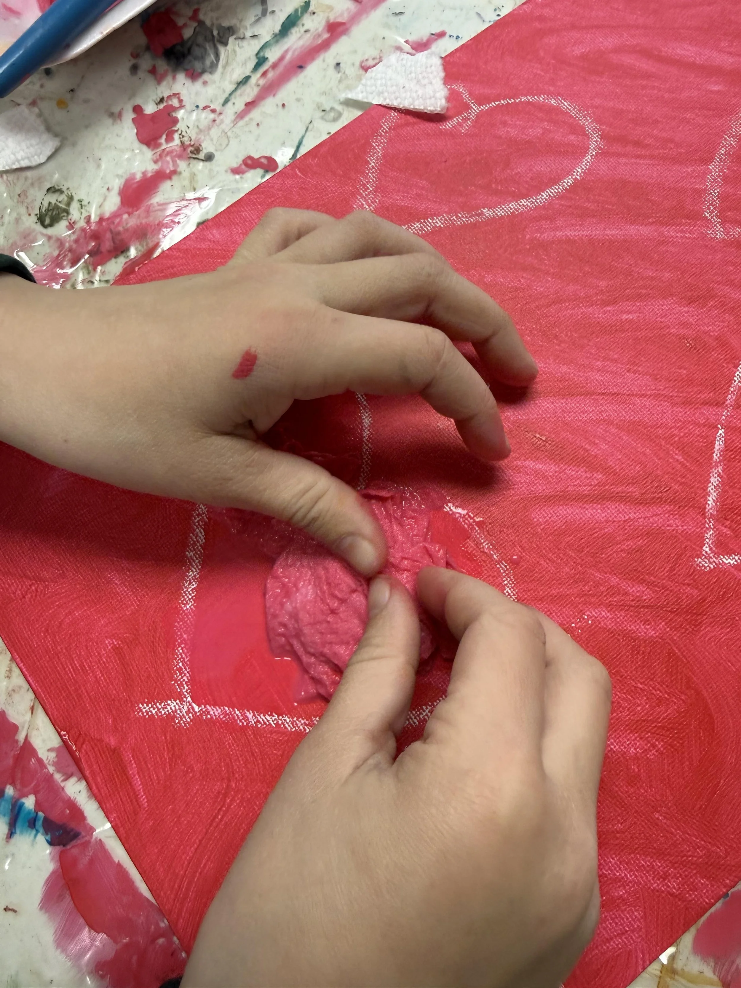 Two hands working on a red canvas or fabric with white heart shapes, surrounded by art supplies, with the hands peeling or tearing a piece of textured pink material.