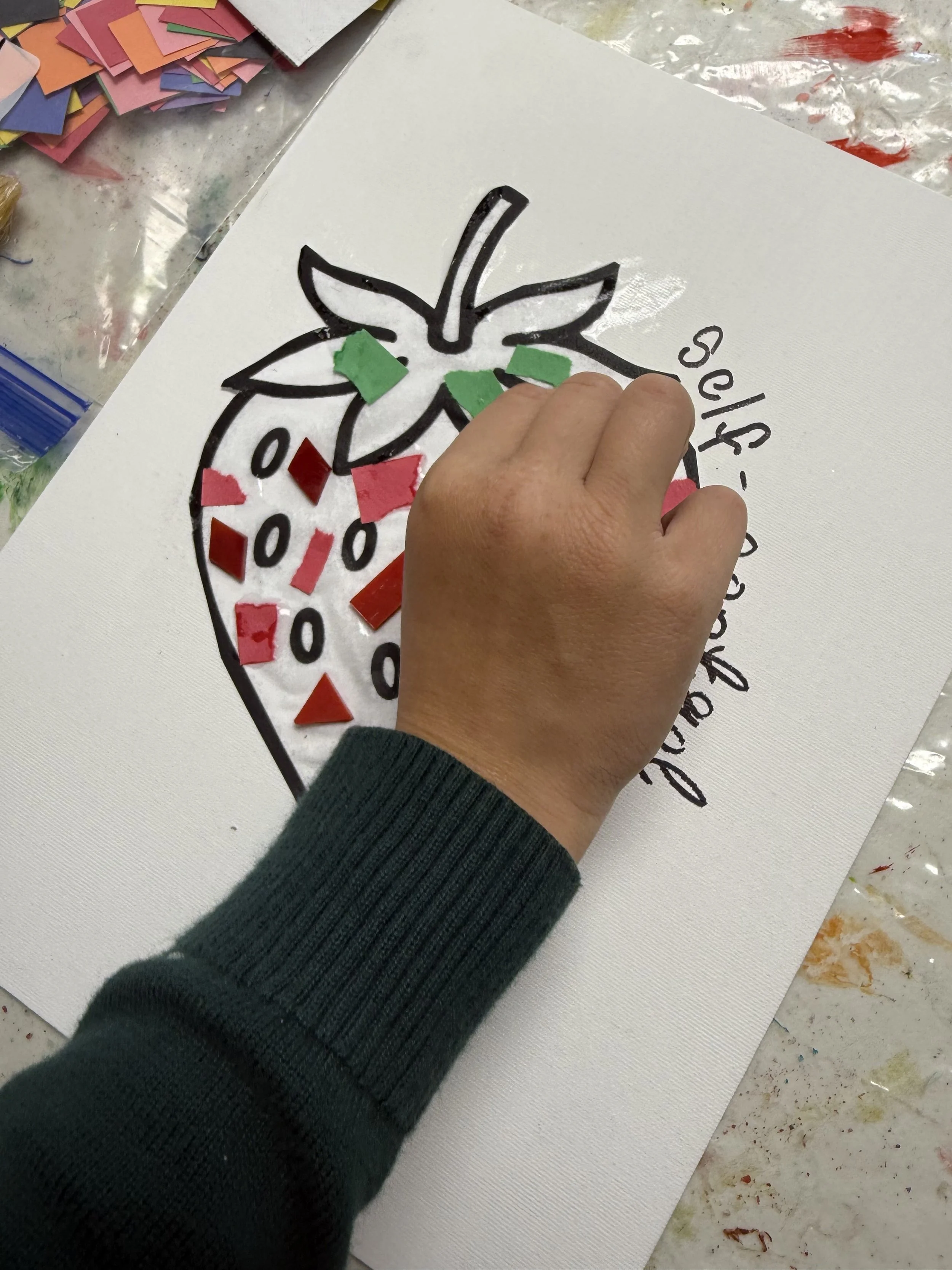 A person gluing small red and green pieces onto a worksheet with a outlined strawberry drawing, beside the partially colored strawberry, the words 'Strawberry' and some colorful paper scraps on the table.