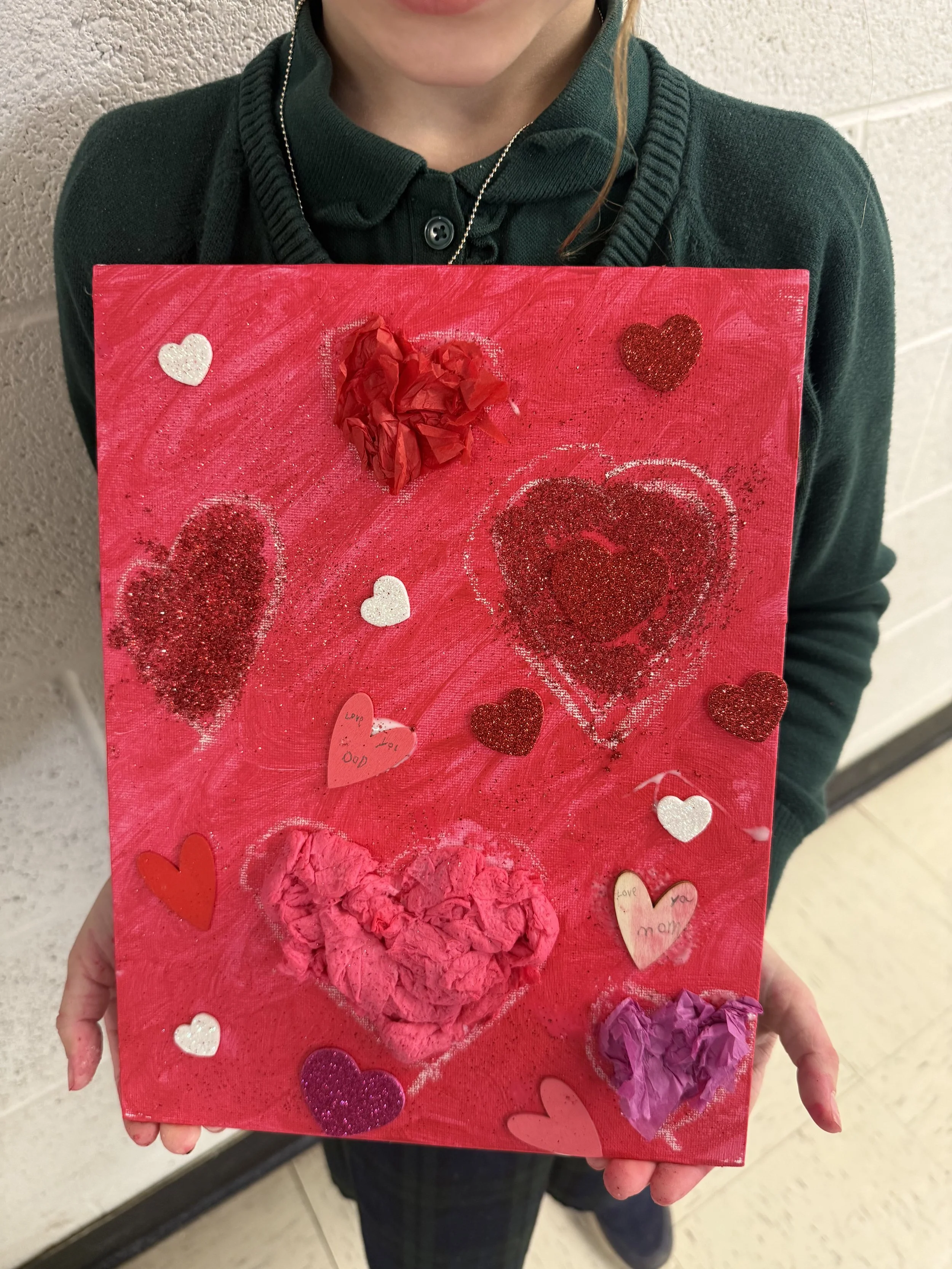A child in a dark green sweater holding a decorated Valentine's Day craft board with hearts and messages.