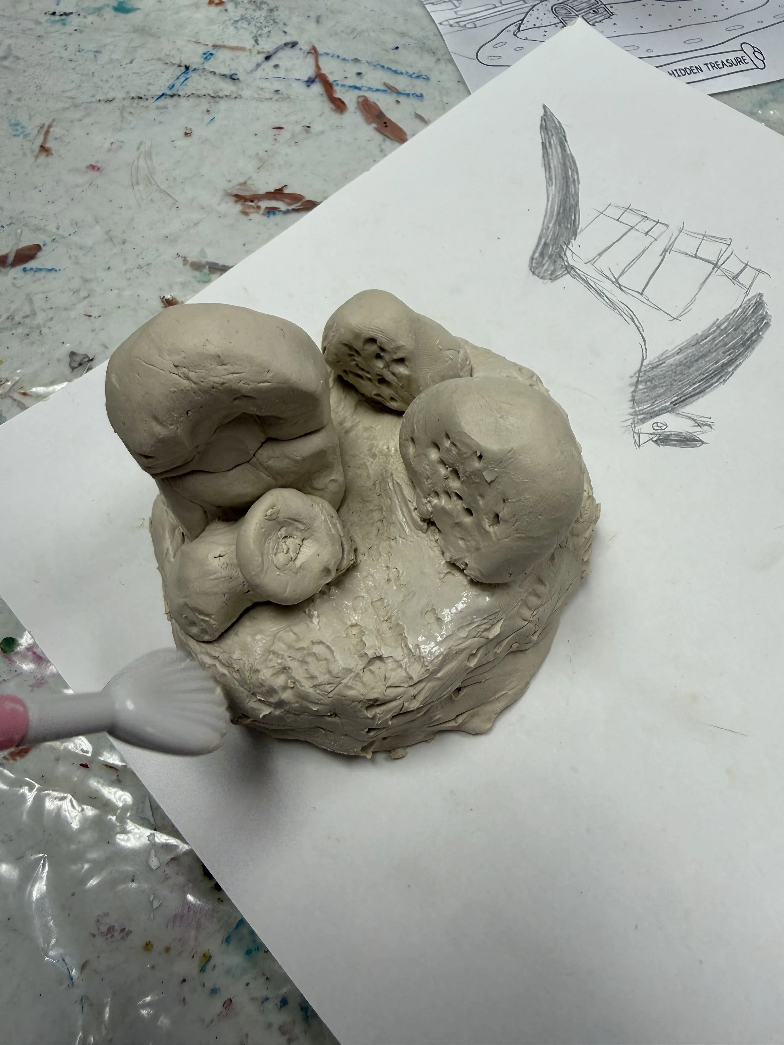 A clay sculpture of mushrooms on a piece of paper with a pencil drawing of a mushroom and a chair. The sculpture shows different mushroom caps and stems with details in the clay.