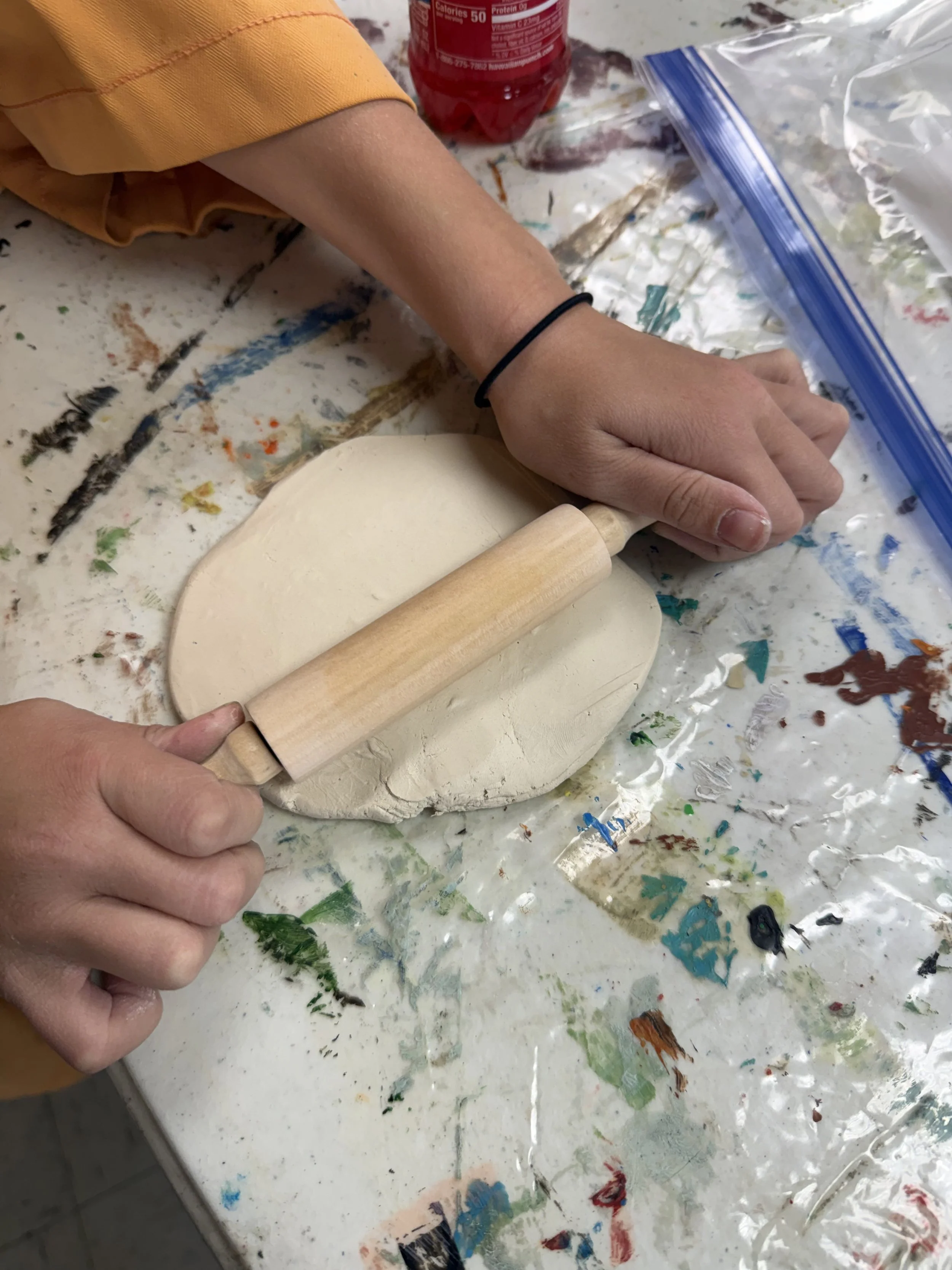 Person rolling out clay with a rolling pin on a table covered in paint splatters, with a plastic bag and a red soda bottle nearby.
