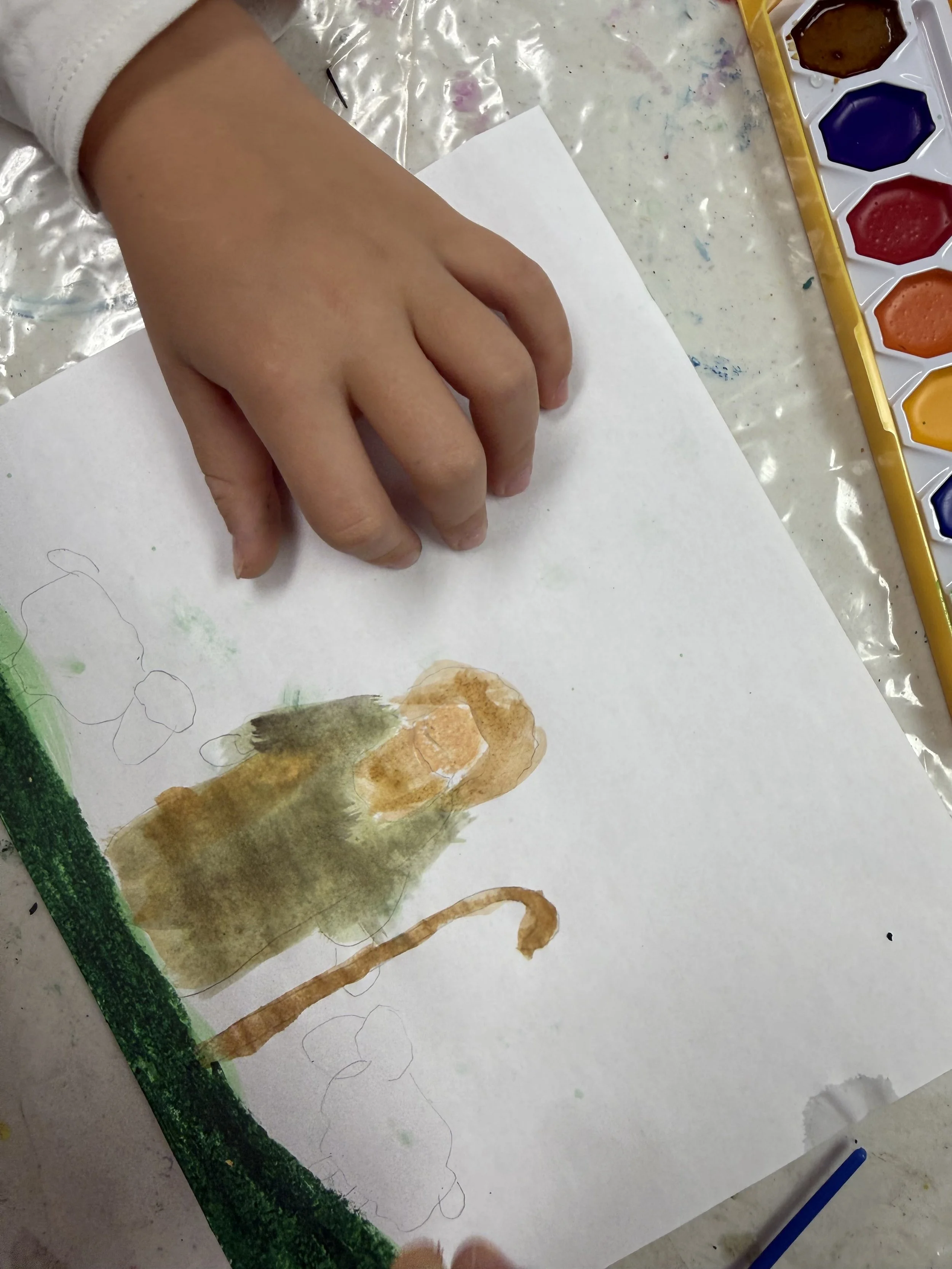 Child's hand working on a watercolor painting of a monkey on white paper, with watercolor paints visible nearby.