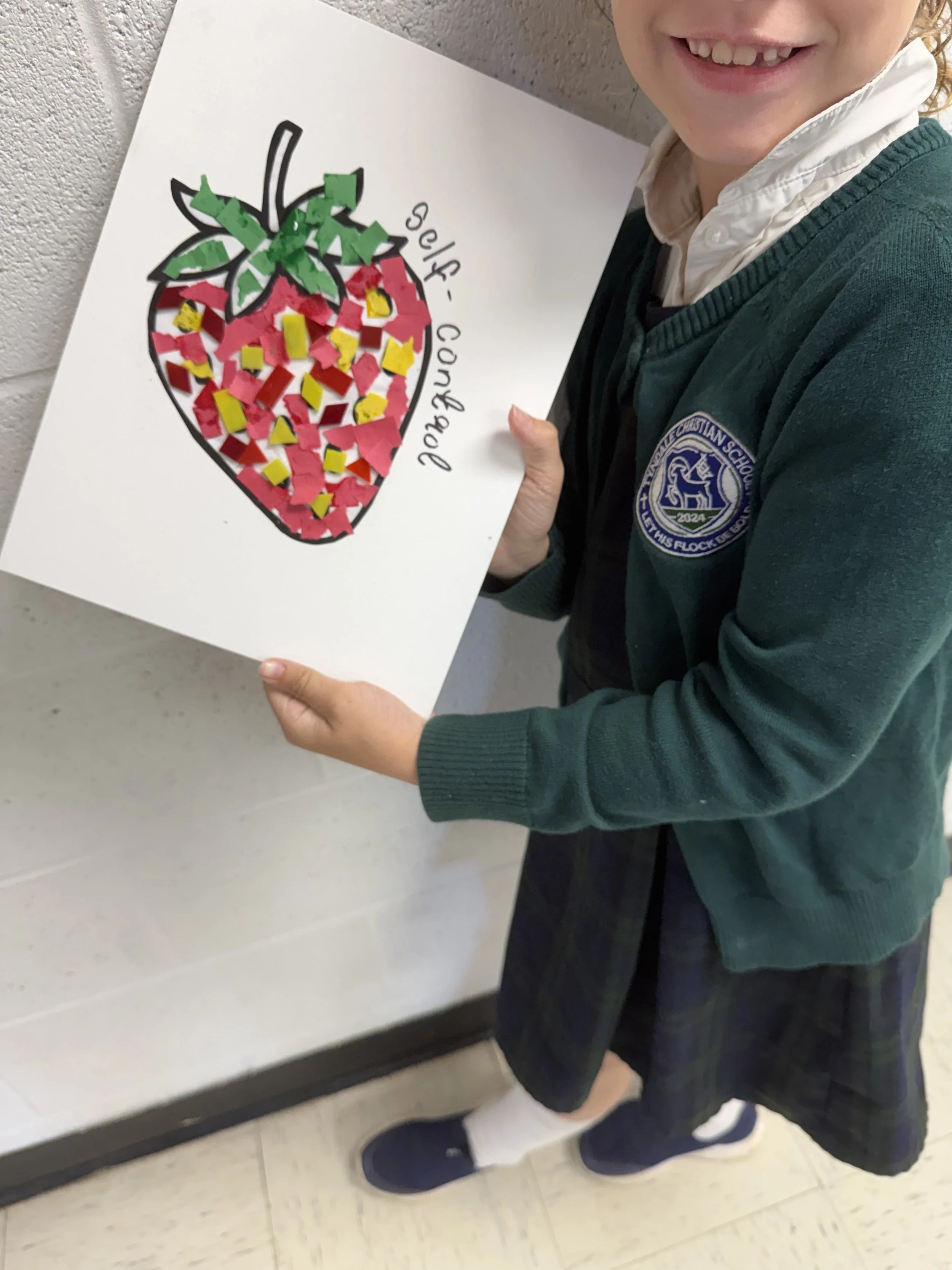 A student in a school uniform holding a collage of a strawberry created with paper and stickers, with the words 'self-control' written on it.