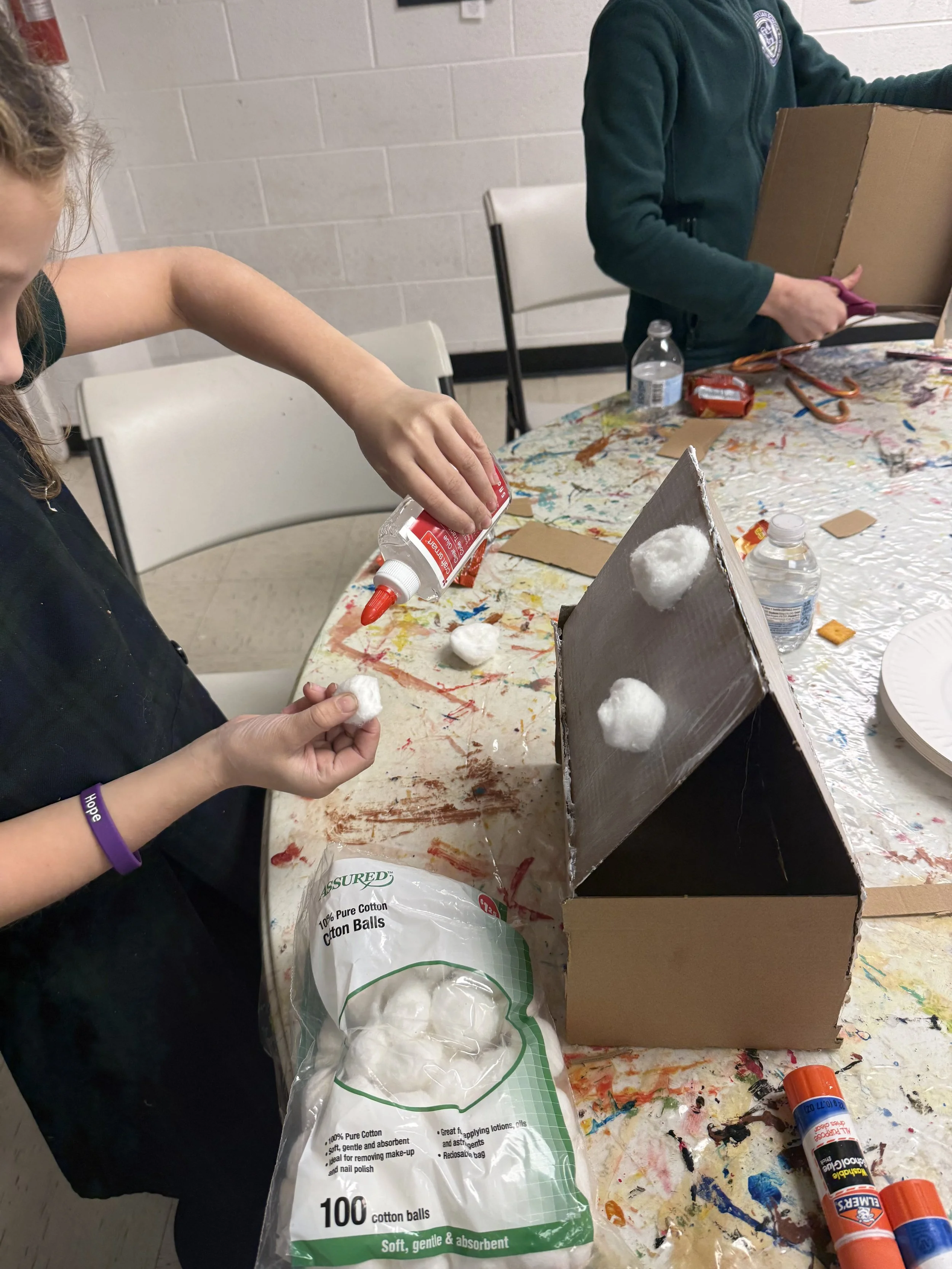 A girl working on a craft project, placing cotton balls onto a cardboard structure, with art supplies and cotton balls on the table.