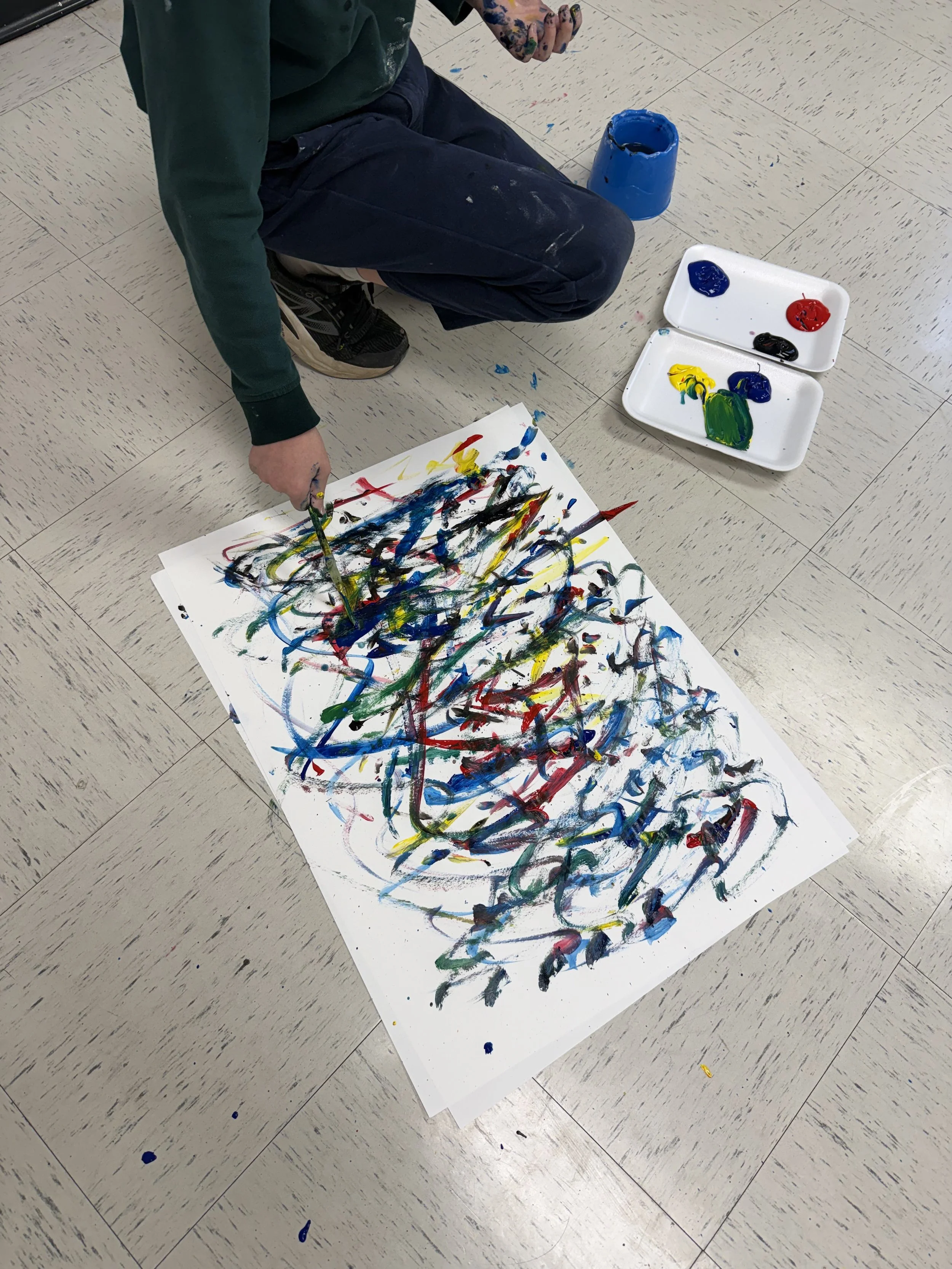 Child kneeling on beige tiled floor, creating an abstract painting with colorful paint splatters and strokes on a large sheet of paper. Paint containers and trays with red, blue, yellow, green, and black paint nearby.