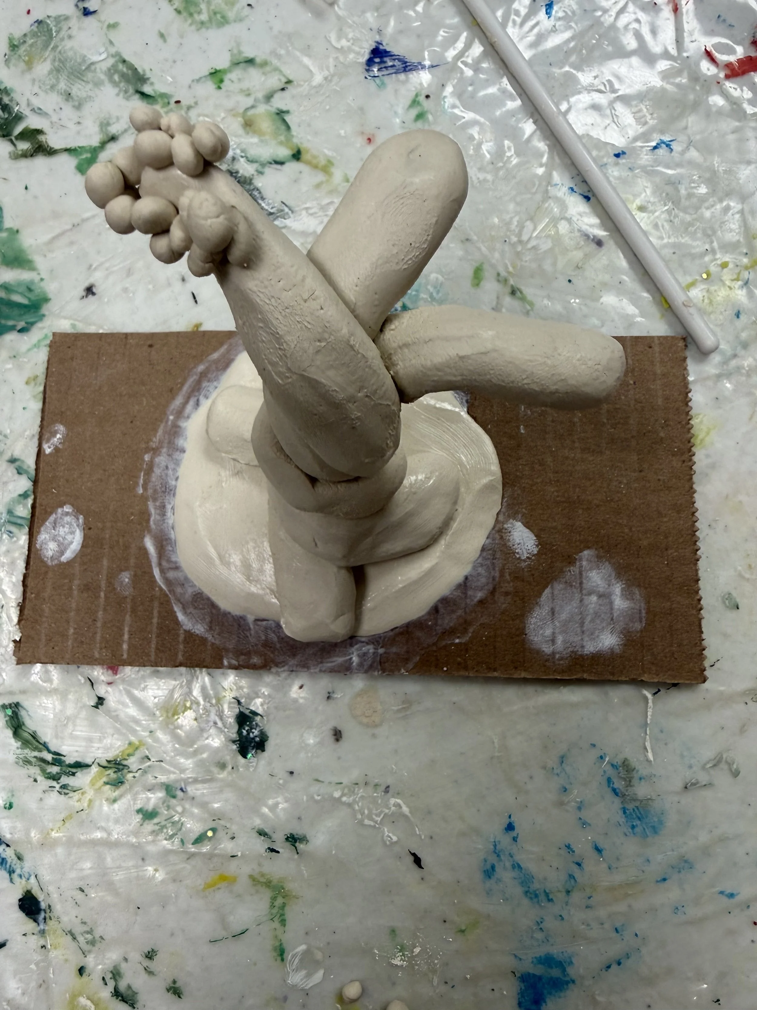 A sculpture made of clay resembling a cartoonish, abstract figure with multiple rounded shapes and limbs, mounted on a piece of cardboard with a painted base.