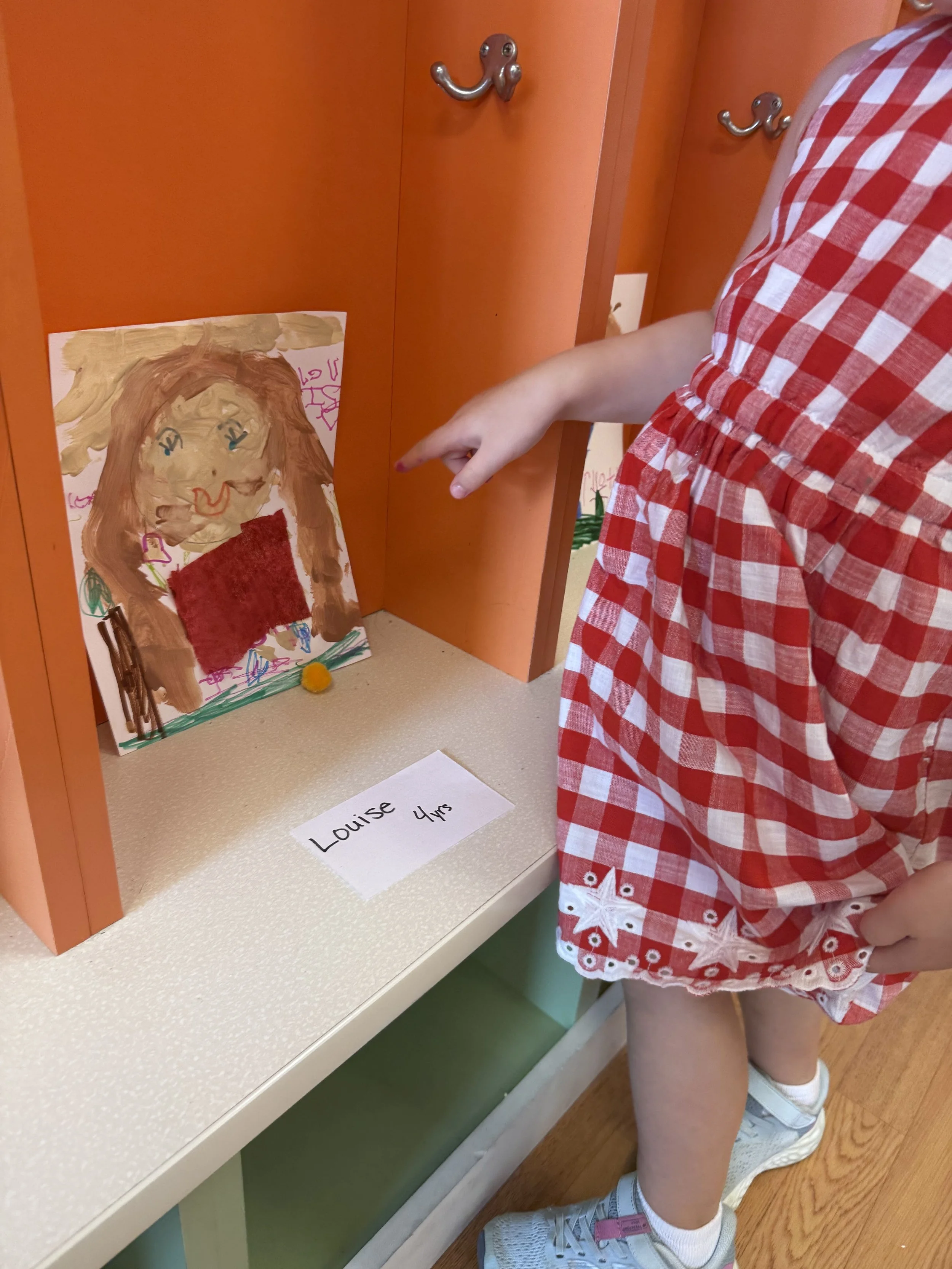 Child in a red and white checkered dress pointing at a hand-painted picture of a woman with long brown hair and a red dress, displayed on a shelf with a small yellow pom-pom at the base. A white card with black text reads "Louise 4 yrs."