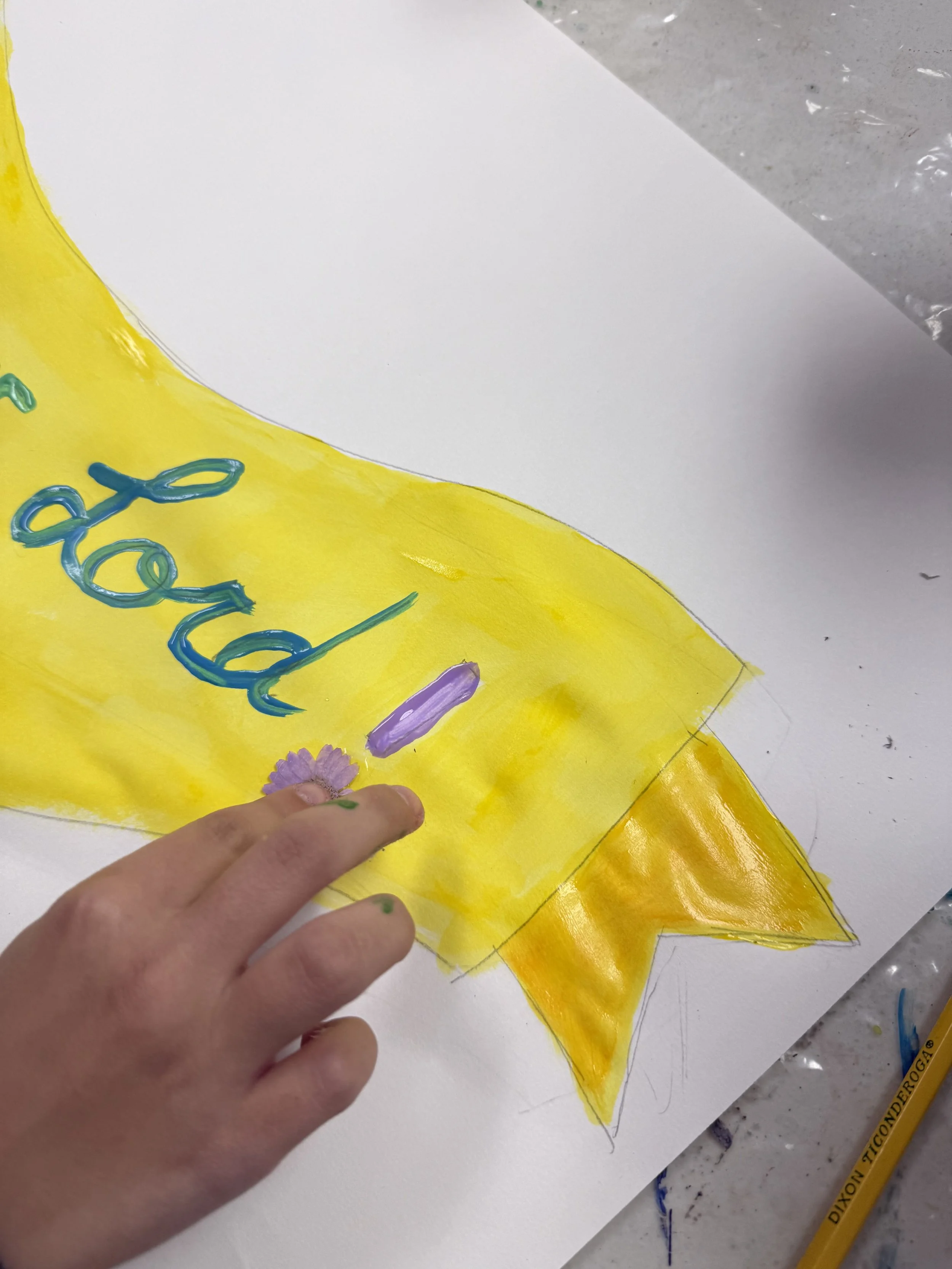 Colorful handmade peace sign with the words 'peace' in blue cursive, a purple flower, and a small purple object on yellow background, with a hand in the foreground.