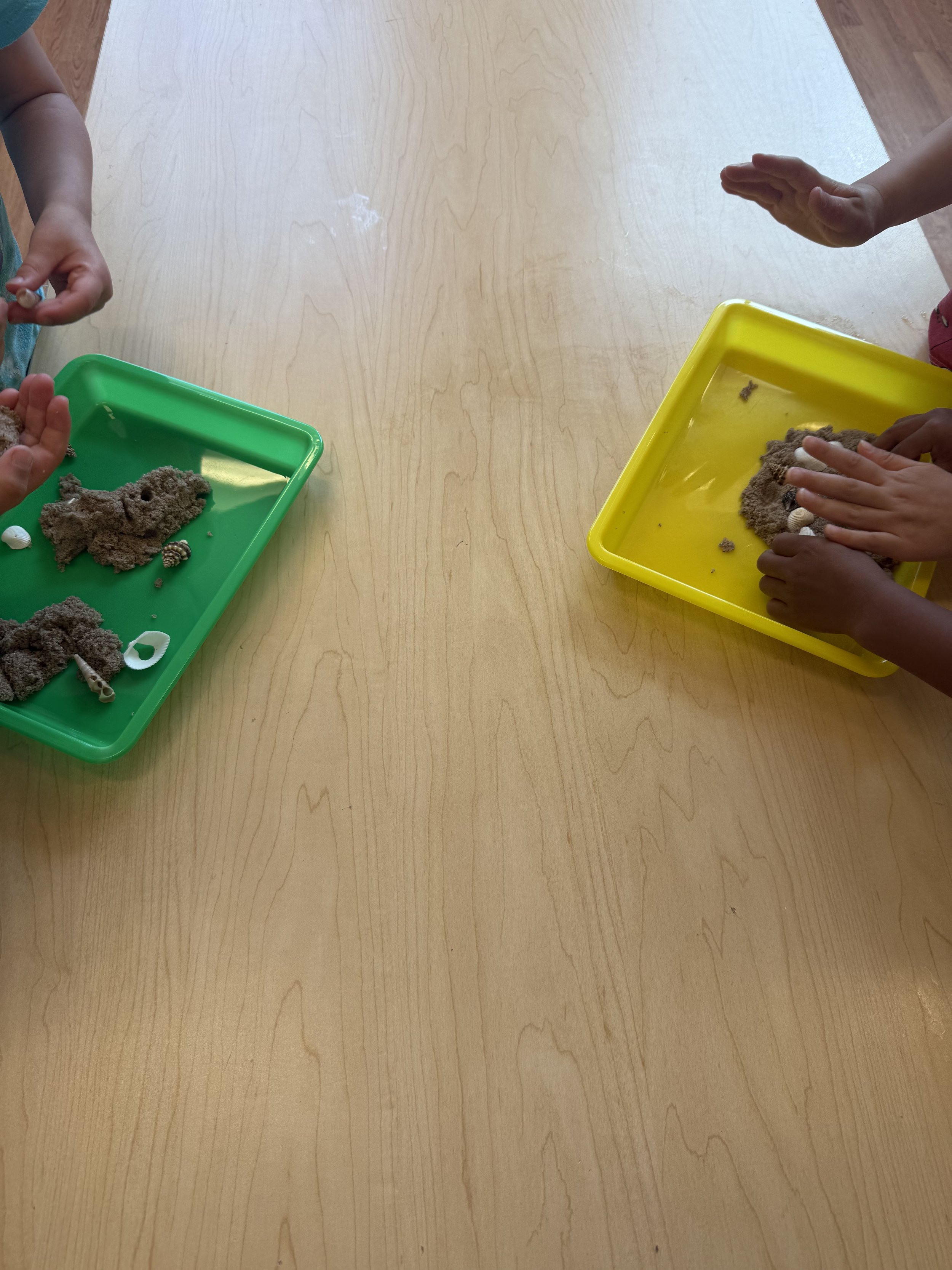 Children playing with brown, dough-like material in yellow and green trays on a wooden table.