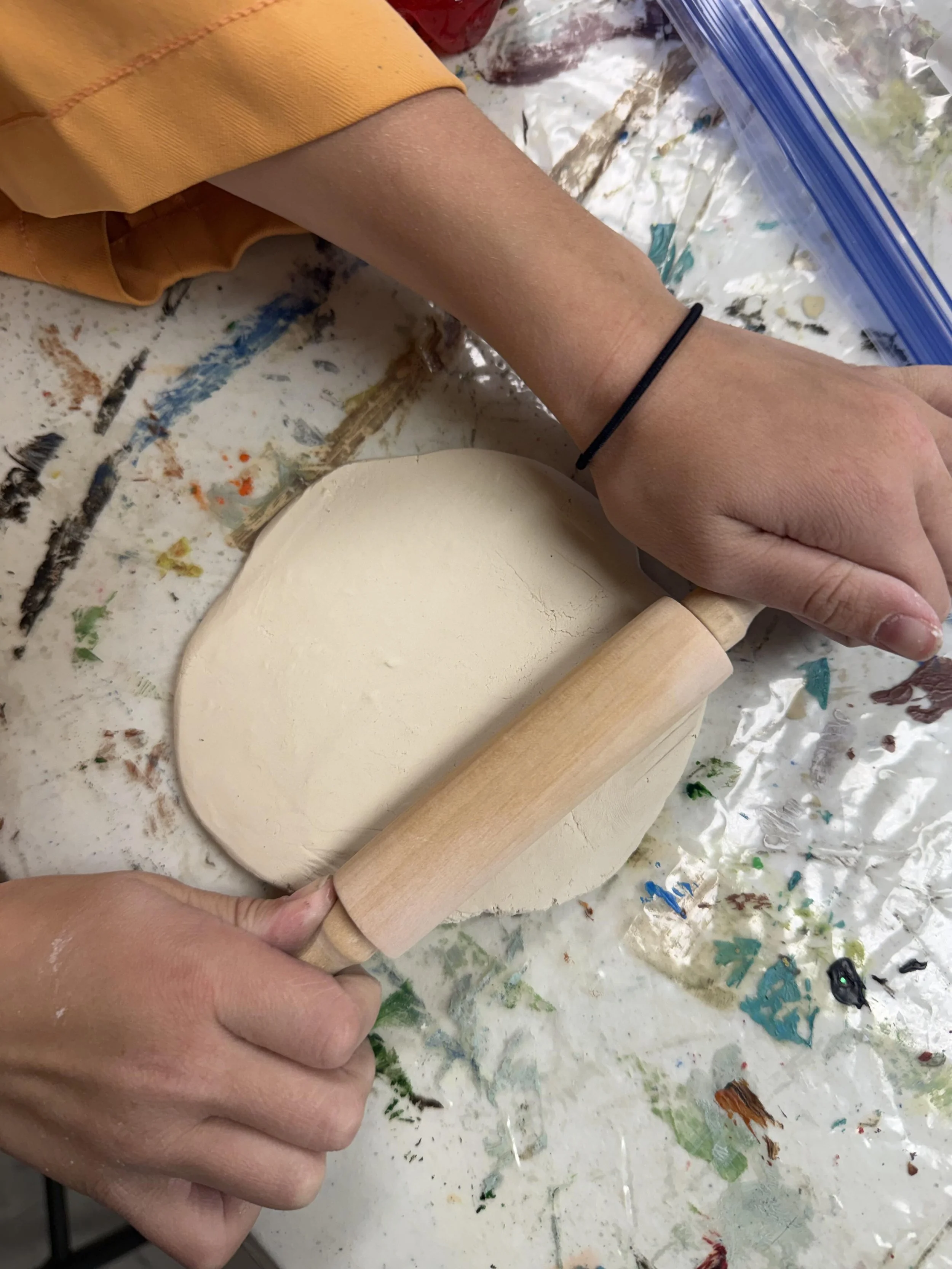 Person using a rolling pin to flatten a piece of dough on a cluttered, paint-splattered work surface.