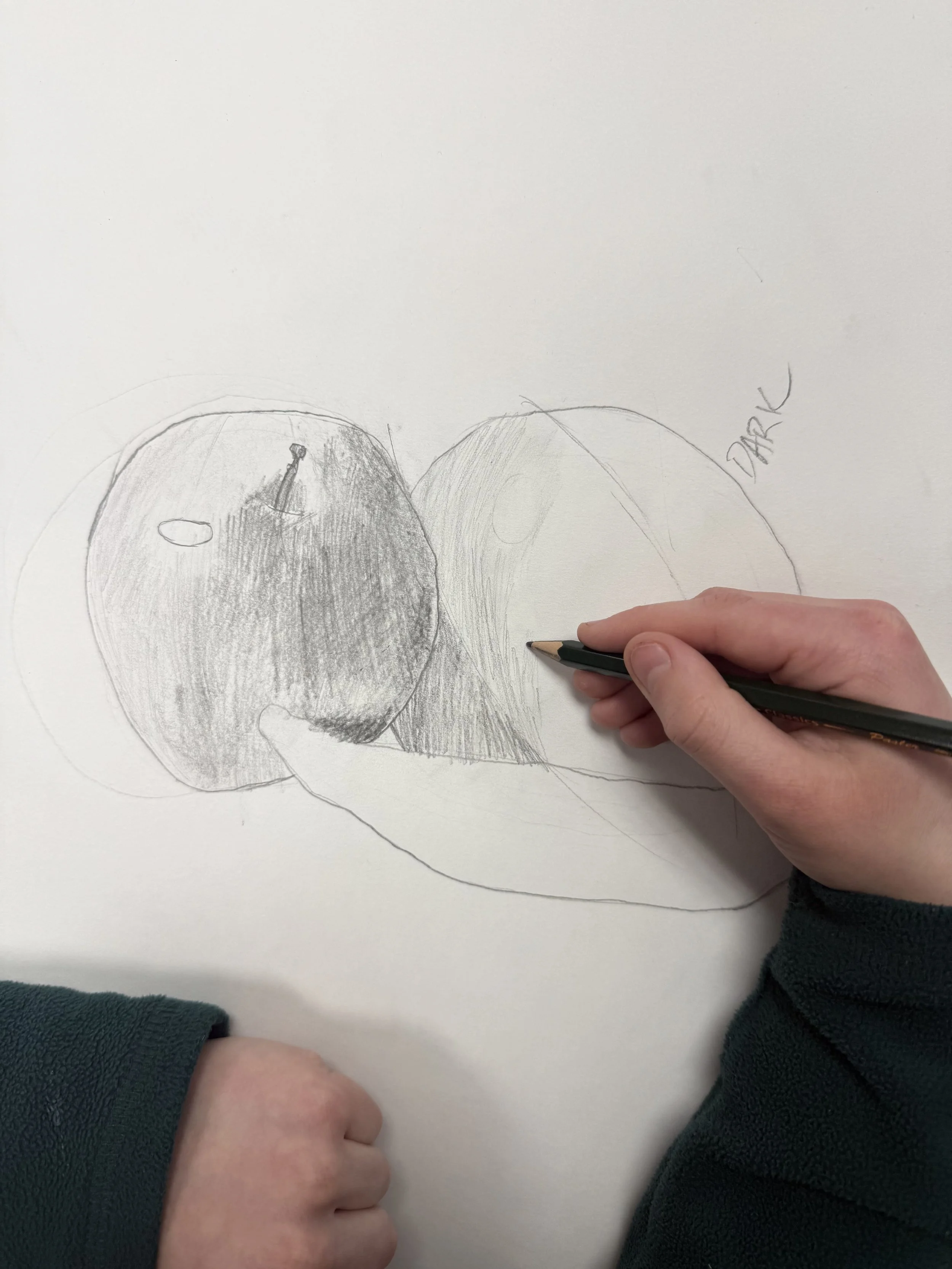 Hand sketching a fruit, possibly an apple or peach, on white paper with a pencil, with the word 'Dark' written nearby.